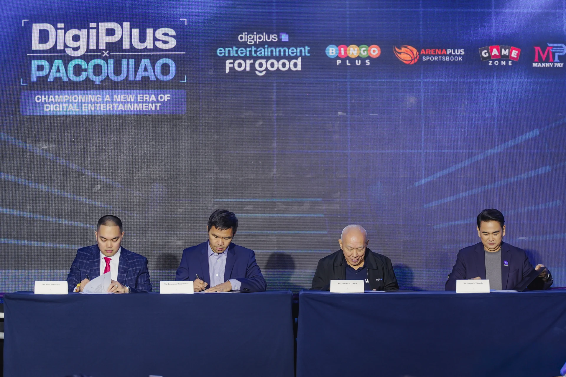 Four men in suits sign documents at a long table during a formal signing event, backdrop shows DigiPlus and logos like Pacquiao and Bingo Plus.