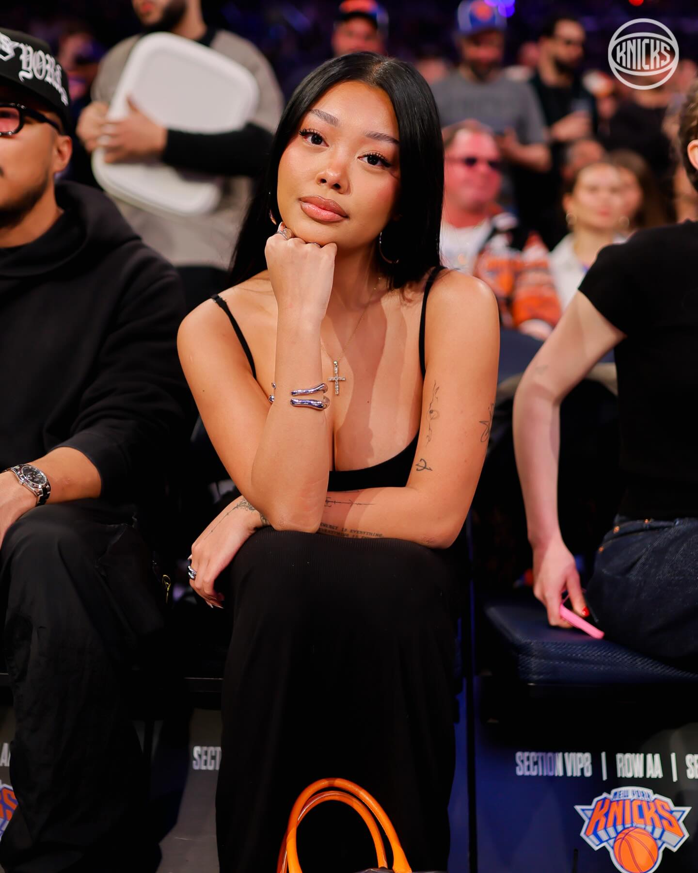 669830026_18581787613010608_4026318416287467328_n - the game Young woman with long black hair sits in arena seating, chin resting on her hand, wearing a black spaghetti-strap dress and hoop earrings at a Knicks game.