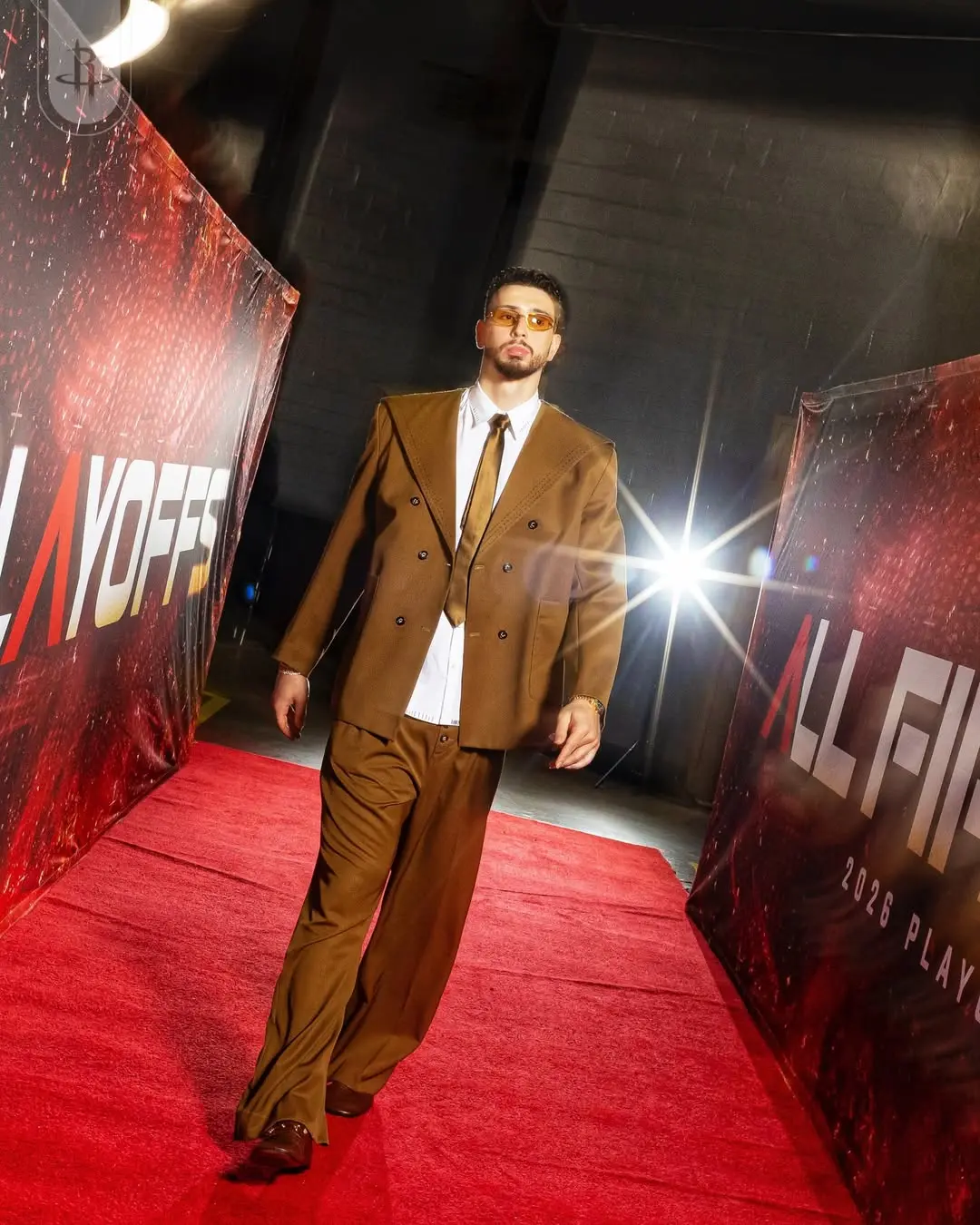 Alperen sengun - the game Man in a brown double-breasted suit walks the red carpet at a fashion event, wearing white shirt, brown tie, and amber-tinted sunglasses.