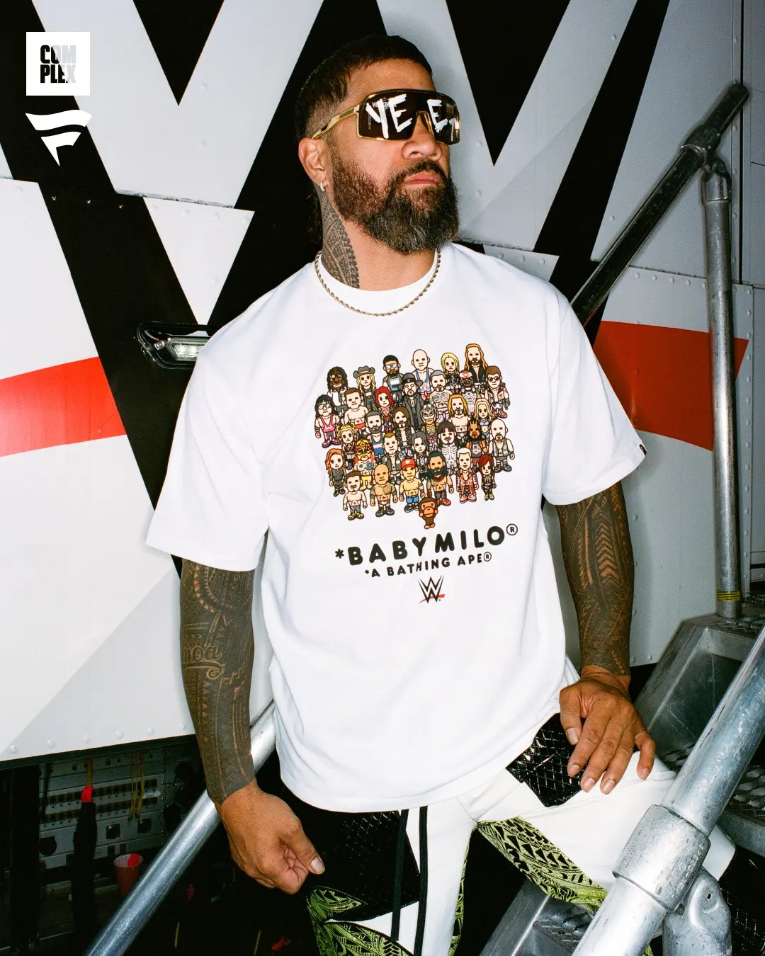 Man with a full beard and sunglasses poses on a metal staircase wearing a white Baby Milo graphic tee with a crowd image and chain necklace.