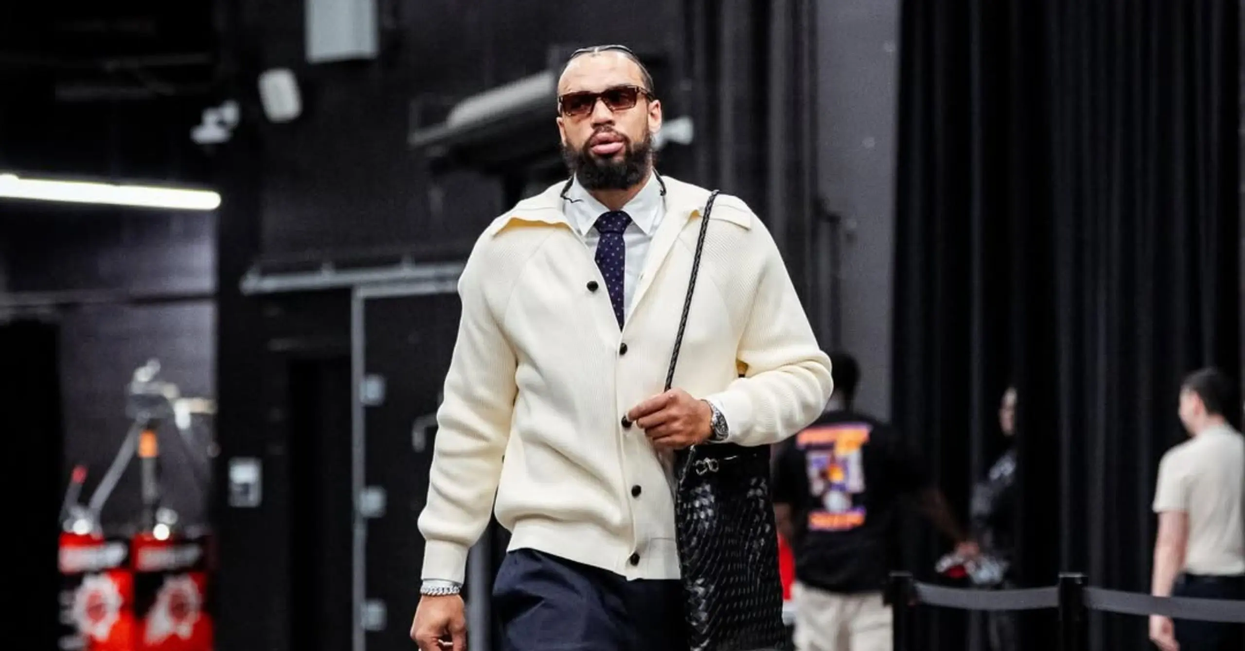 Bearded man in sunglasses wearing a cream cardigan, white shirt, and tie, walking backstage with a black bag over his shoulder.