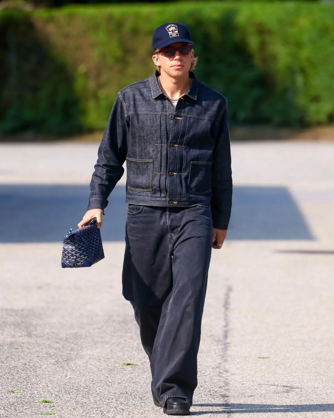 Bistrup - the game Man wearing a dark denim jacket and wide-leg jeans, sunglasses, and a cap, walking on a paved street while holding a blue-patterned clutch bag.