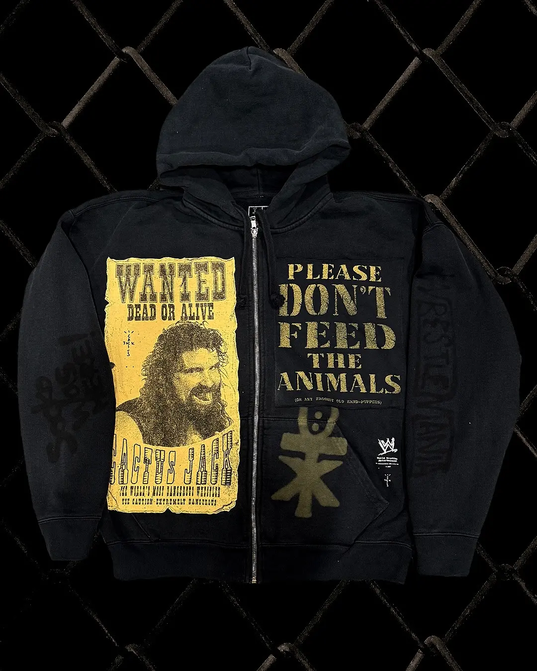 Black zip-up hoodie with a large yellow 'WANTED DEAD OR ALIVE' graphic on the left and bold 'PLEASE DON'T FEED THE ANIMALS' text on the right, set against a dark chain-link fence background.