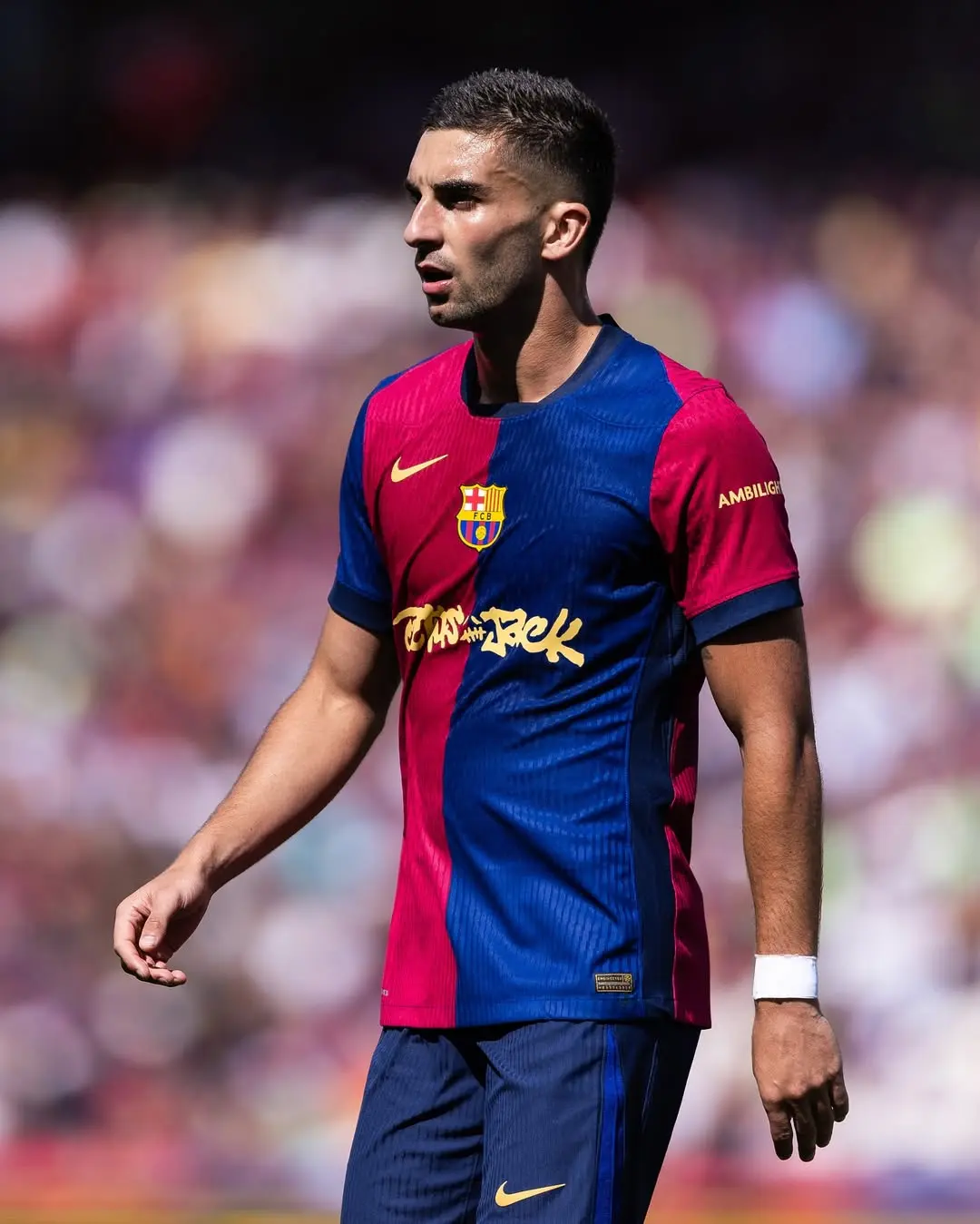 Footballer in Barcelona kit (blue and maroon) walking on the pitch during a match, crowd blurred in background.