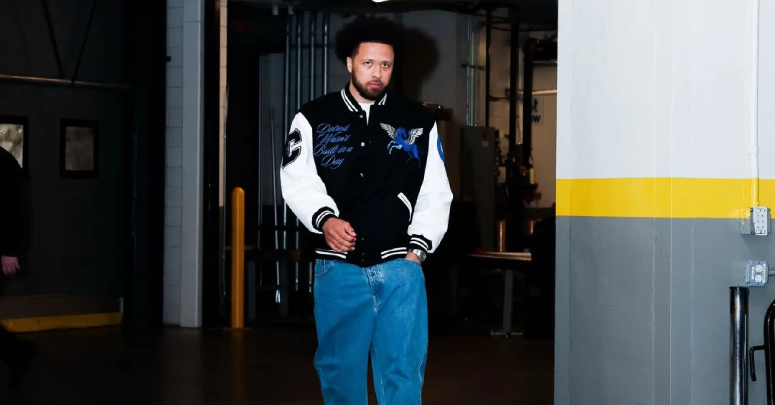 Man with a beard wearing a black-and-white varsity jacket with blue wing design stands in a dim warehouse corridor.