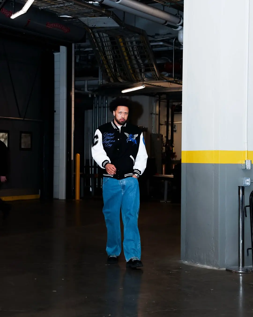 Man with curly hair in a black-and-white varsity jacket and blue jeans walks down a dim, industrial corridor.