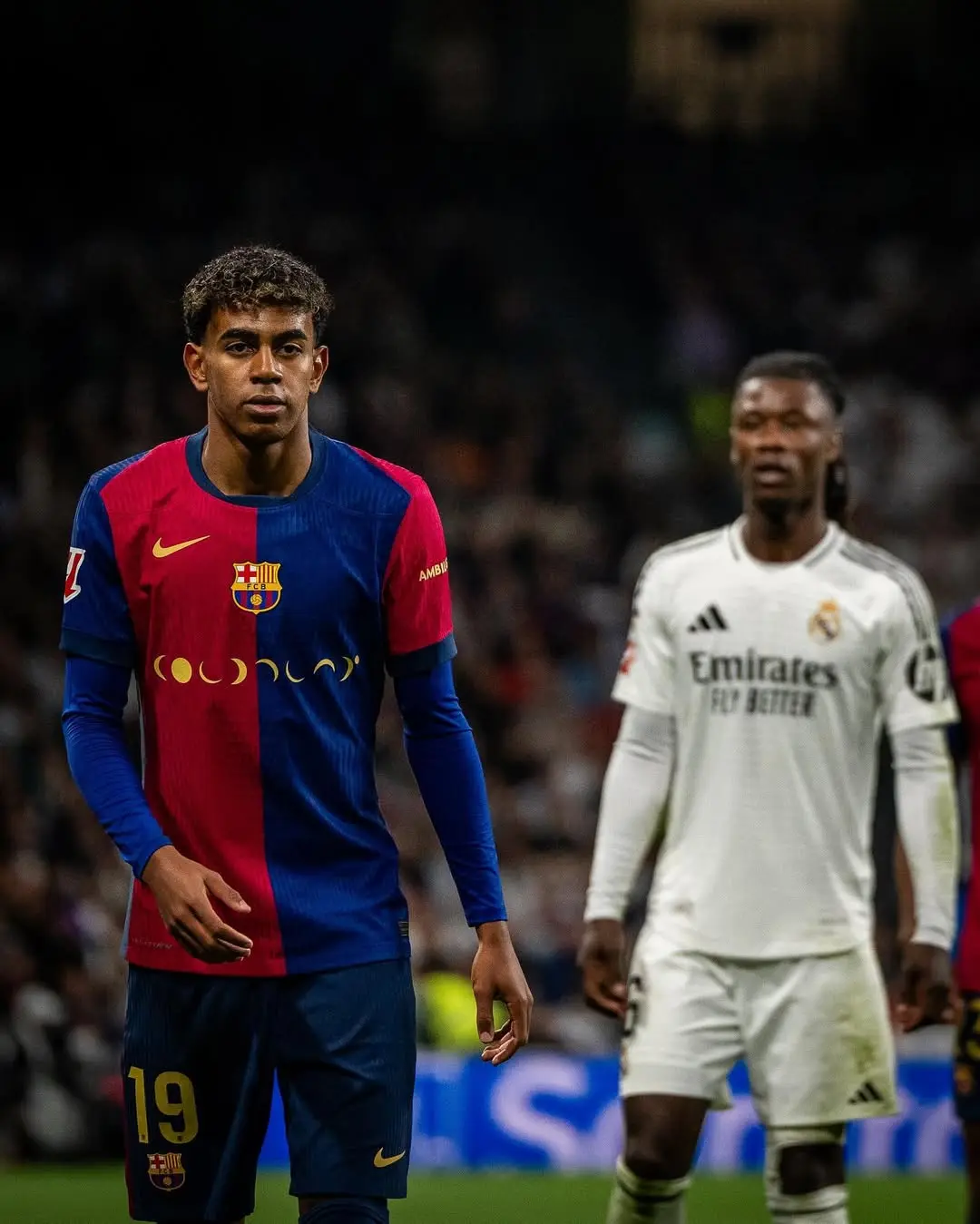 Young footballer in a Barcelona red-and-blue kit walking on the pitch; Real Madrid player in white in the background.