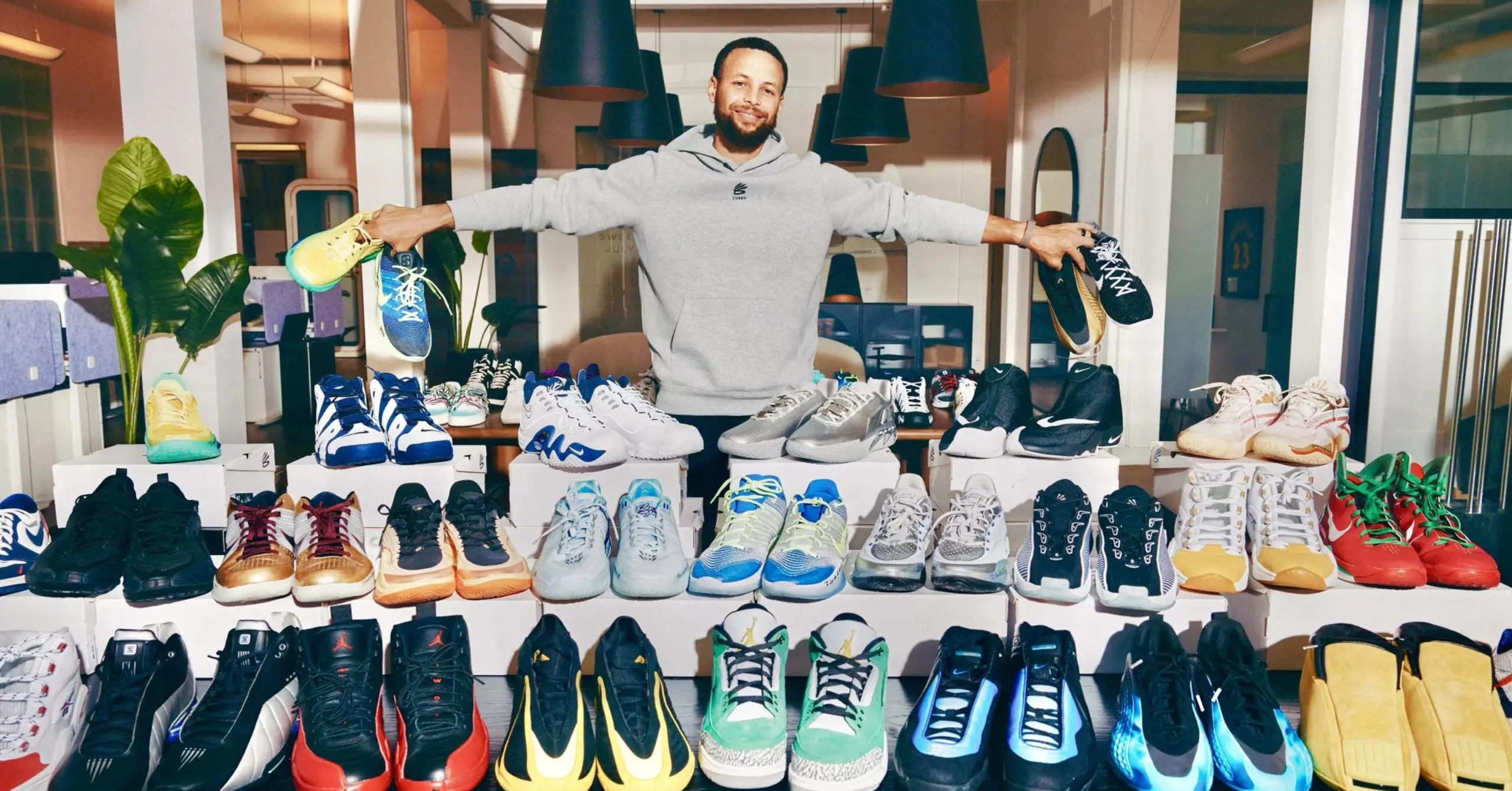 Smiling man in a gray hoodie stands behind a display of colorful sneakers, holding a shoe in each hand.