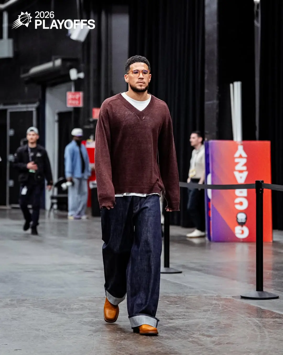 Devin booker - the game Male model walking backstage at the 2026 Playoffs event, wearing a maroon V-neck sweater, white shirt, wide dark jeans, and tan boots.