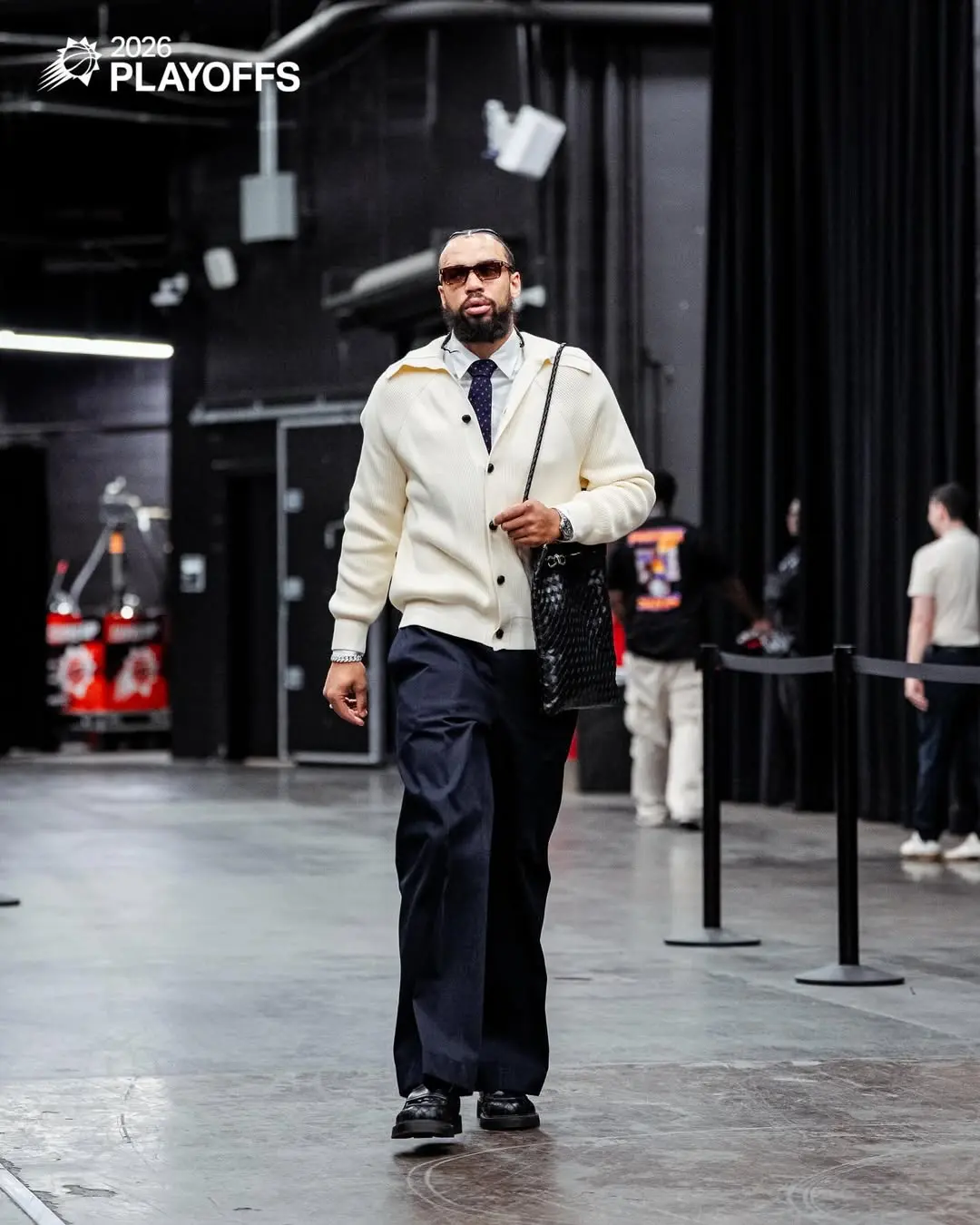 Dillon brooks - the game Man wearing a cream cardigan, navy pants, tie, and sunglasses walks backstage at the 2026 Playoffs, carrying a black textured bag.