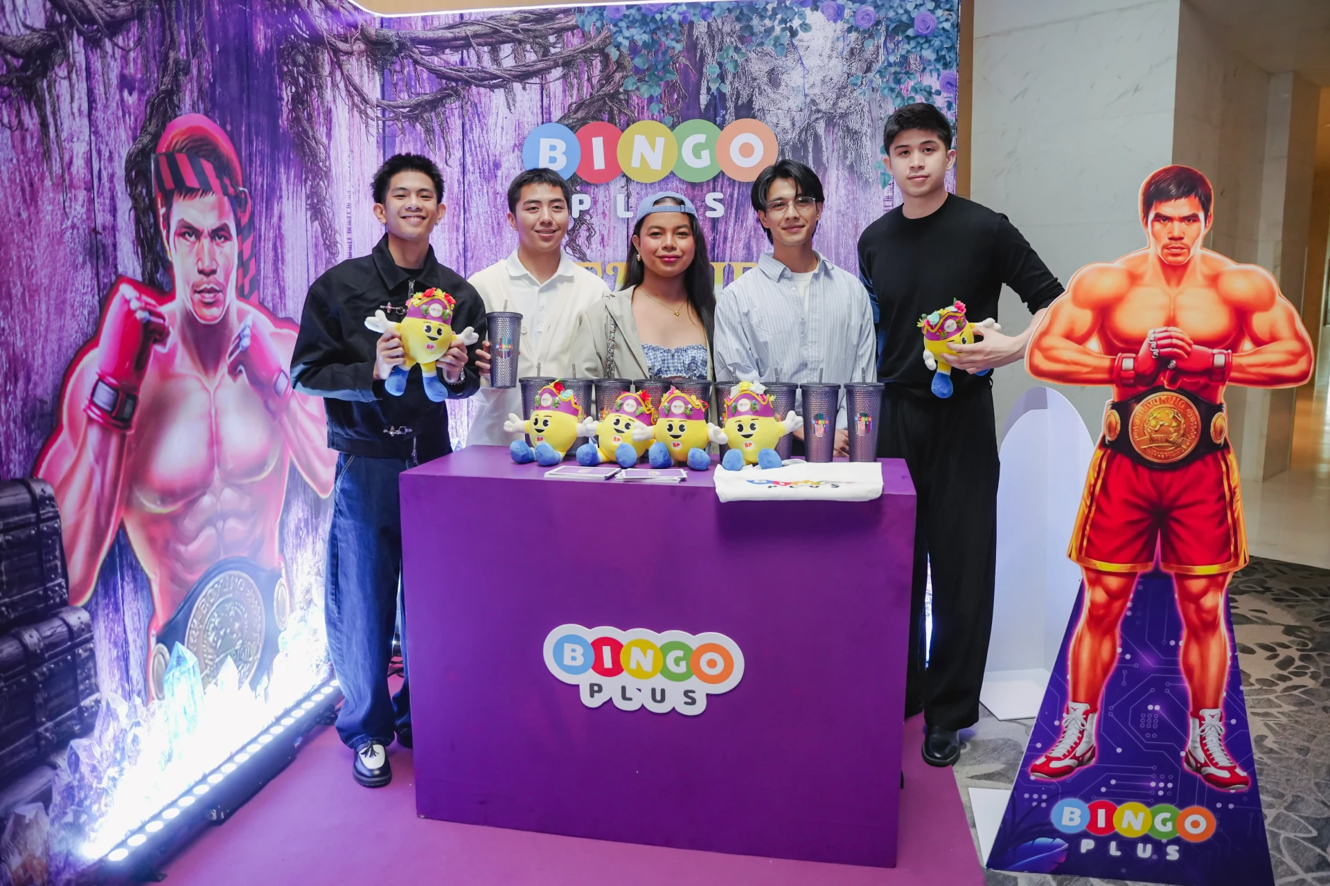 Group of six people posing behind a purple Bingo Plus booth with yellow mascot plush toys on the table and cups nearby; large purple backdrop with boxing figures on either side and a 'BINGO PLUS' sign above.