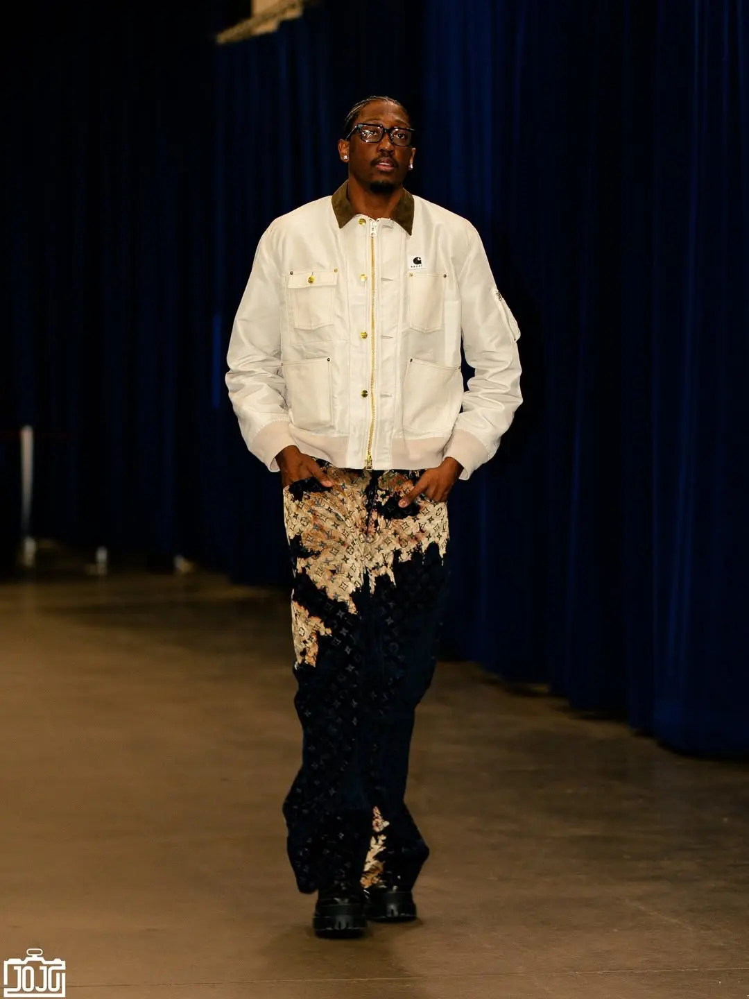 Man wearing a white jacket with brown collar and patterned pants walks down a runway against dark blue curtains, looking ahead.