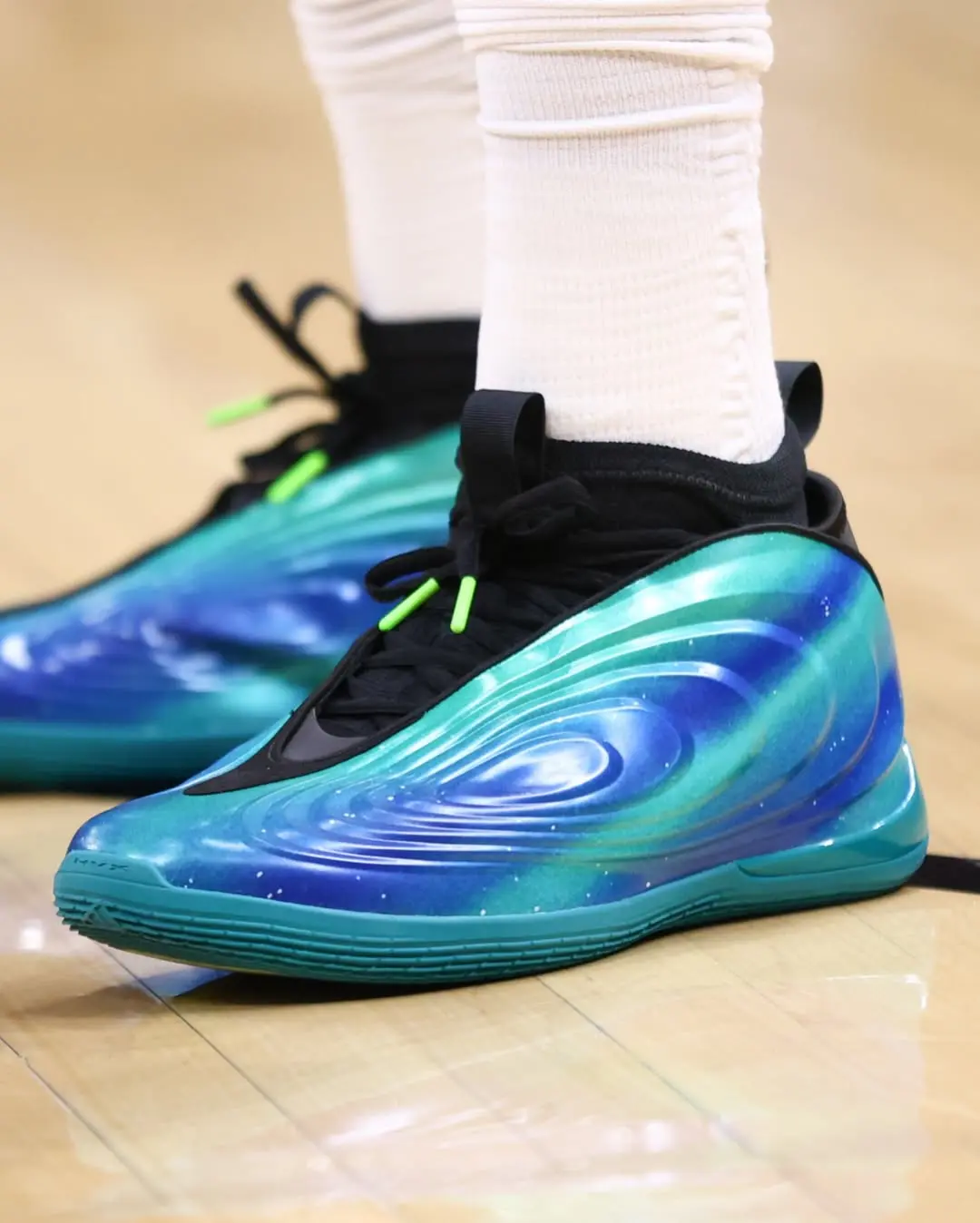 Athletic shoe with a blue-green galaxy pattern and black laces on a wooden floor, white socks visible above the shoe.