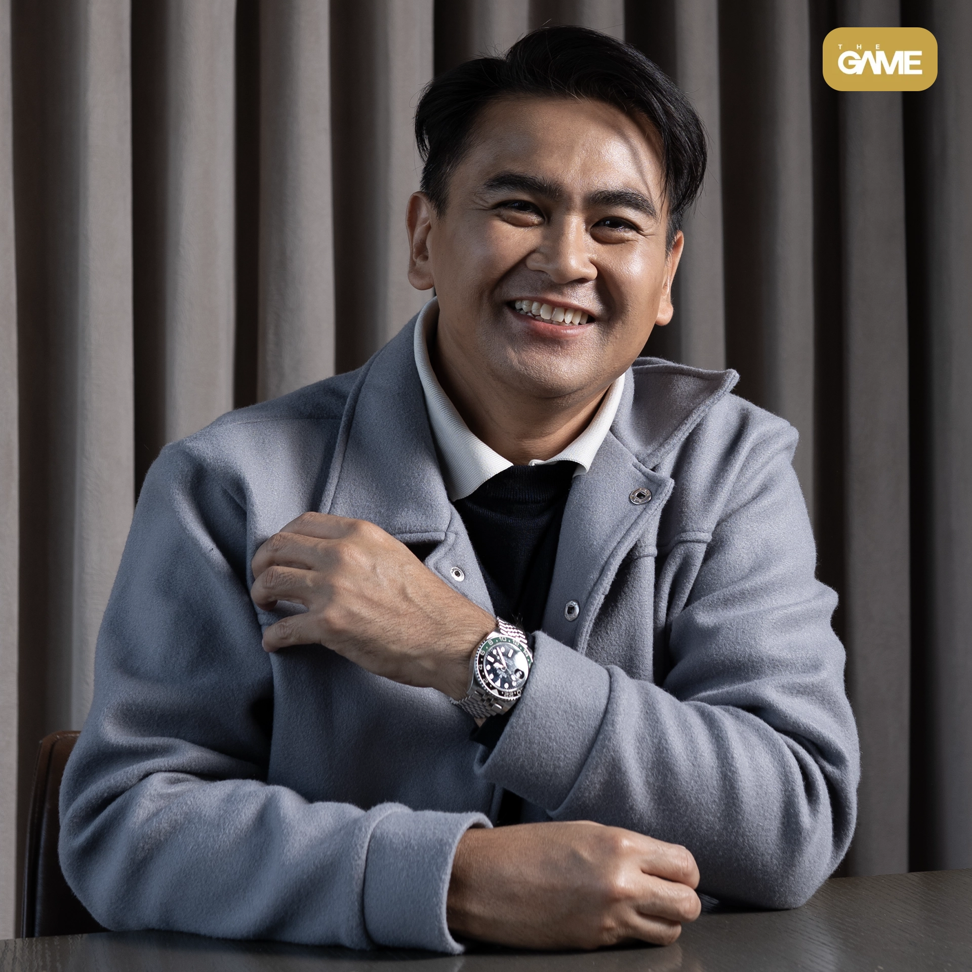 Jasper square - the game Portrait of a smiling man in a gray coat with a silver watch, seated at a table against dark vertical blinds.