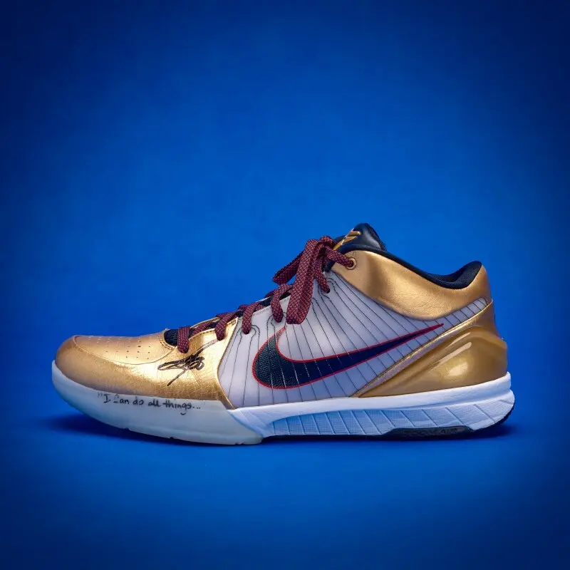 Gold and silver Nike basketball shoe with burgundy laces on a blue background; black swoosh and handwritten quote on the toe.