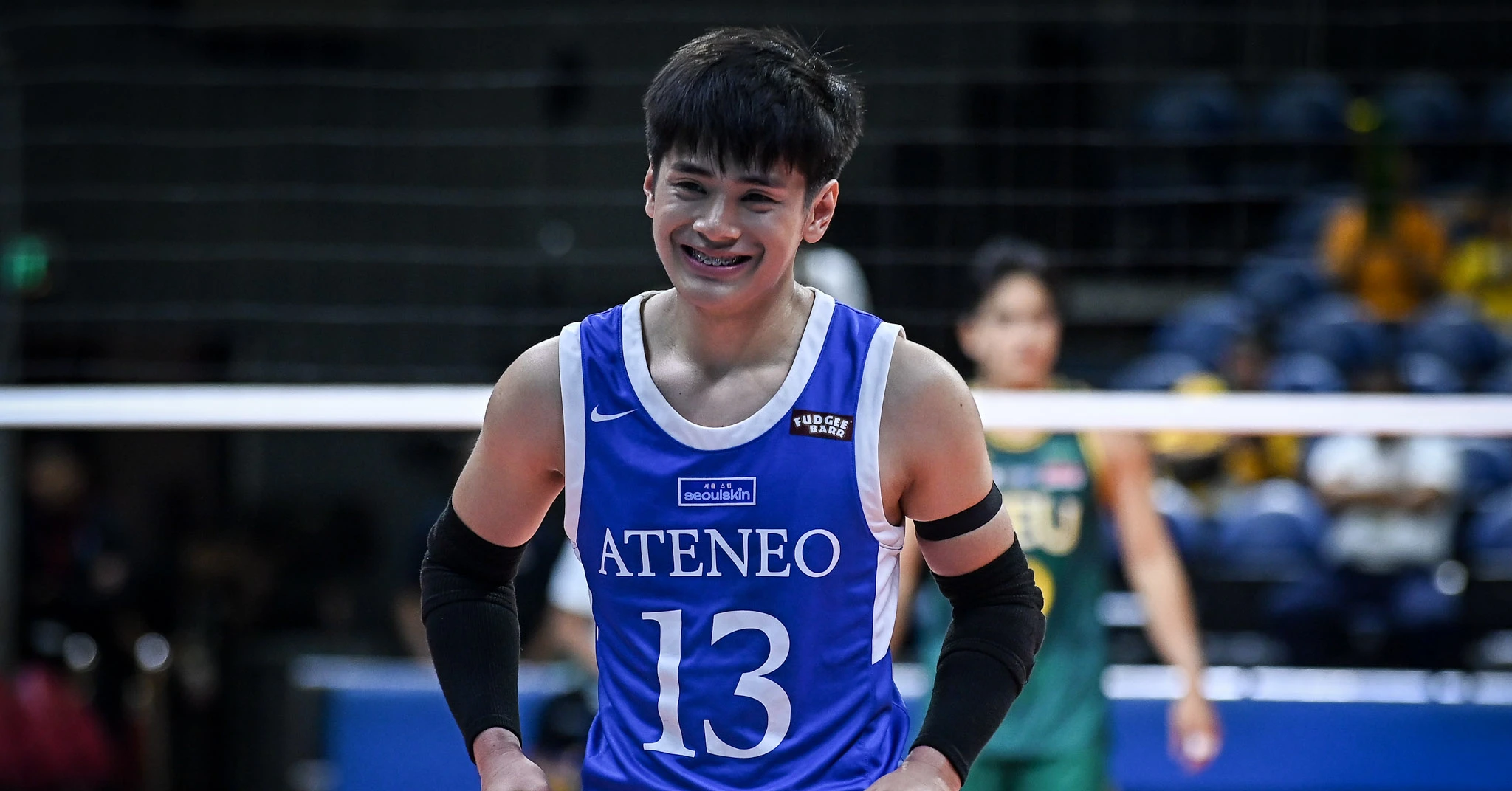 Smiling athlete in a blue Ateneo jersey with number 13 stands on a court during a match, wearing black arm sleeves and braces on her teeth.