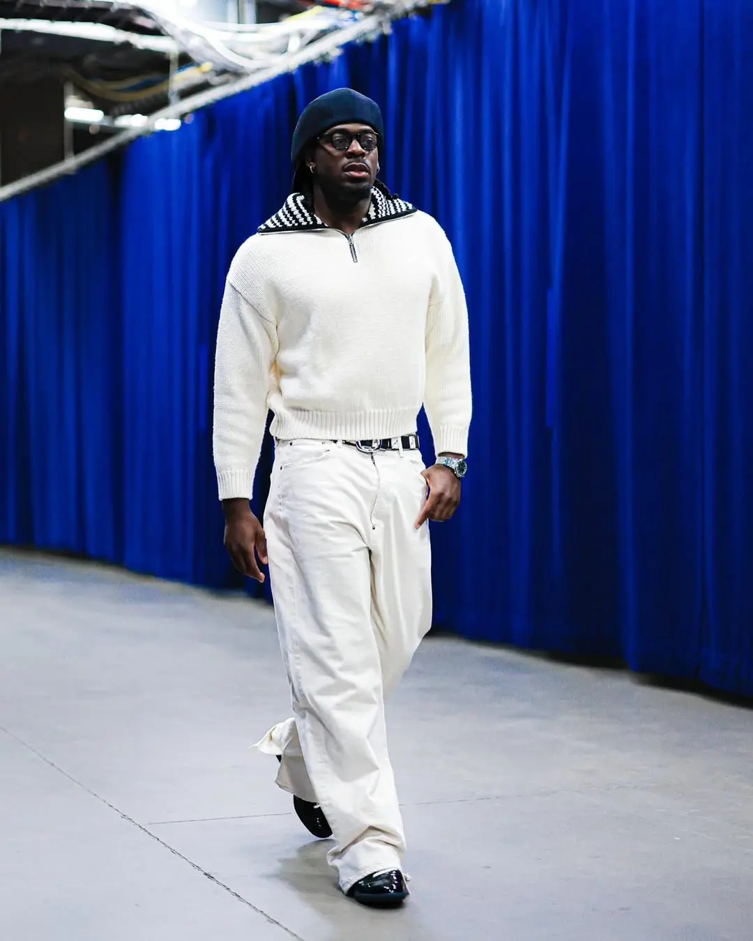 Man in a cream knit sweater, white pants, black beret and sunglasses walks down a hallway with deep blue curtains in the background, posing as a fashion model.