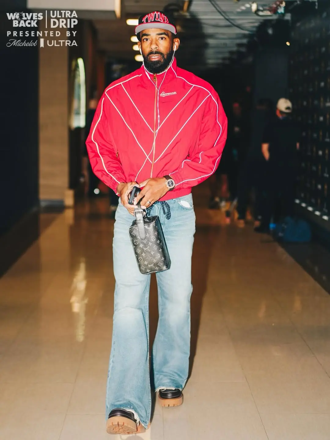 Man in a red track jacket and cap walking in a hallway, holding a small Louis Vuitton bag.