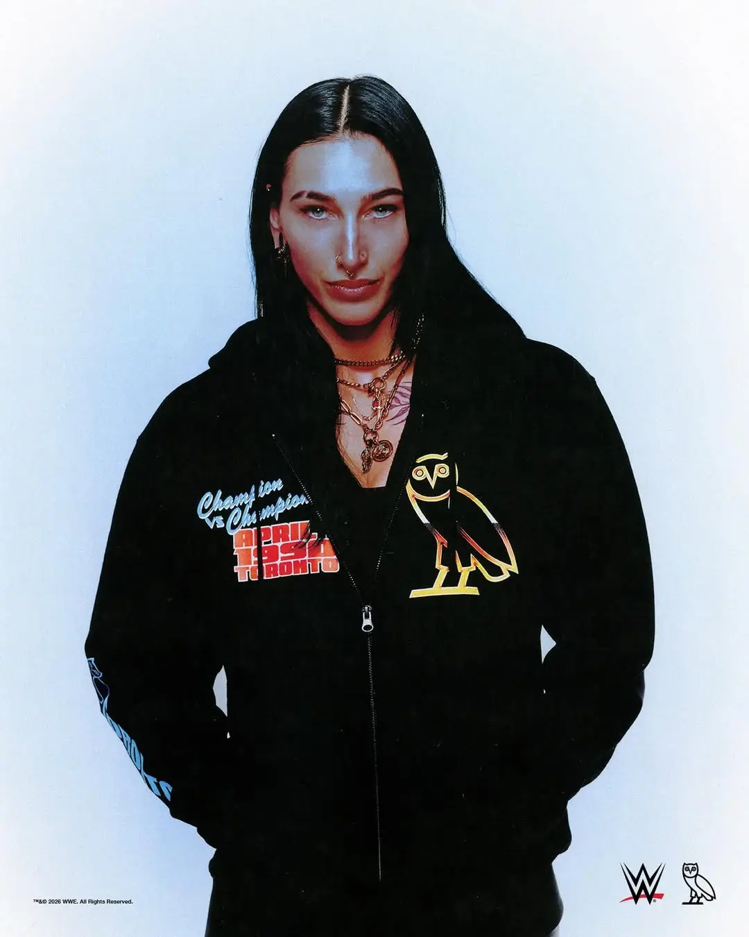 Person with long black hair wearing a black zip-up hoodie with an owl graphic and colorful text on the chest, posing against a pale blue background with hands in pockets.