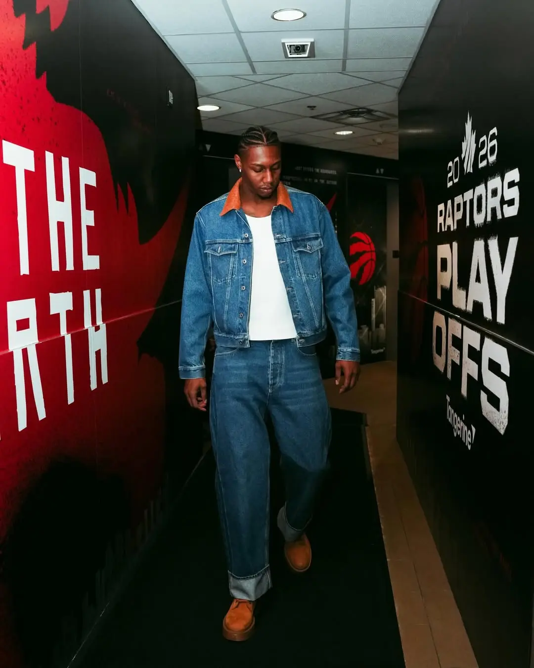 Rj barrett - the game Man in a denim jacket and jeans walks down a hallway with bold Raptors graphics on the walls, brown shoes completing the outfit.