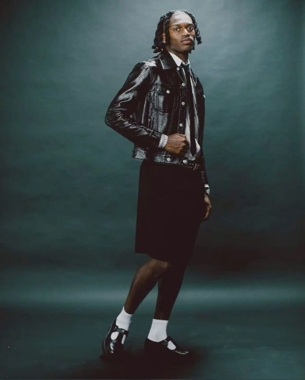 Rafael leao - the game Fashion portrait of a man modeling in a shiny black denim jacket, white shirt, black knee-length shorts, white socks, and black sandals in a studio.