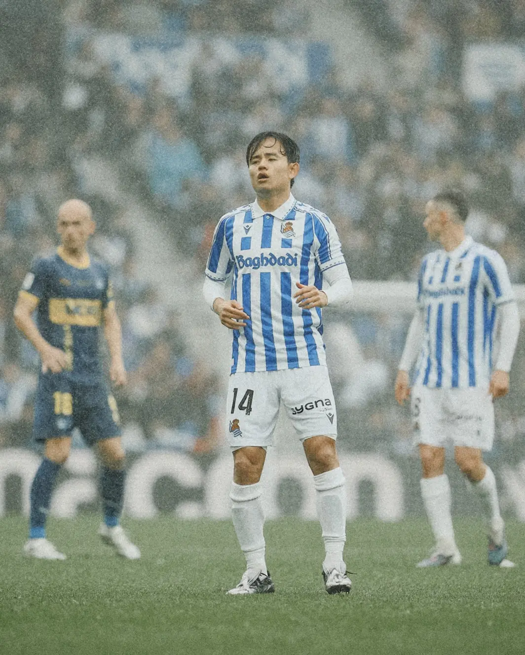 Soccer player in blue-and-white striped kit with number 14 stands on a rain-soaked field during a match.