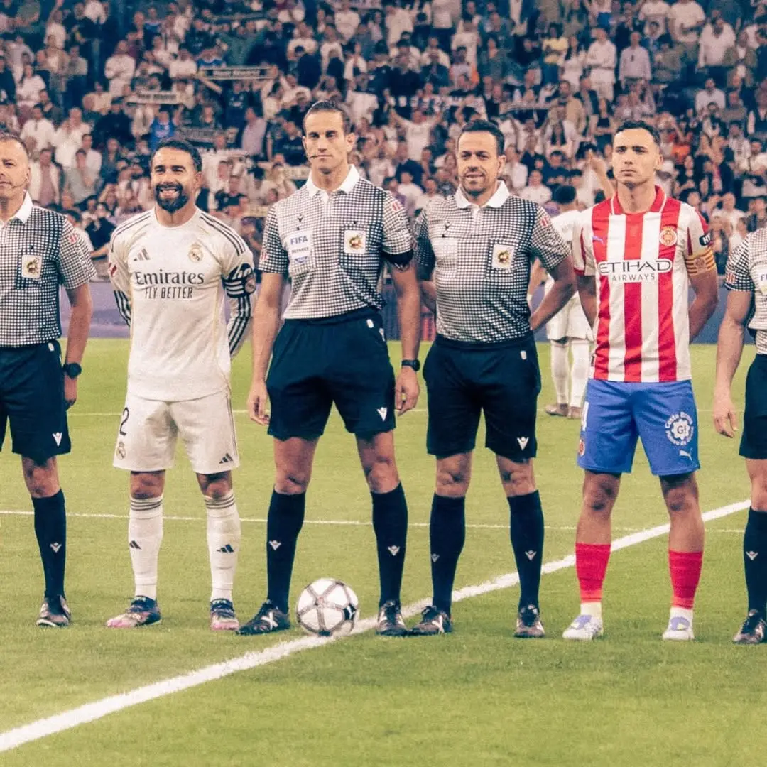 Lineup of players and referees on a soccer field before kickoff, with a ball at their feet and a crowded stands in the background.