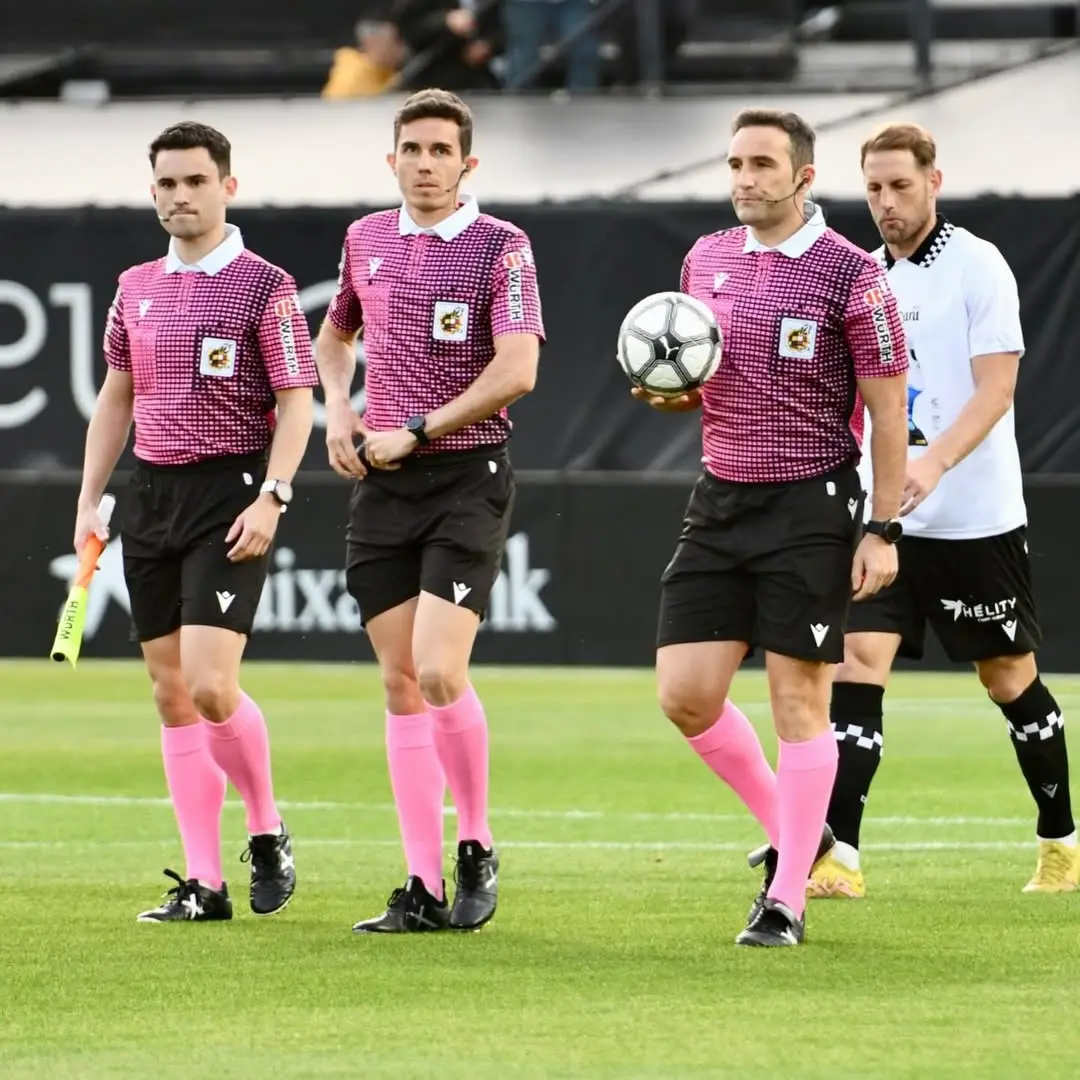 Four soccer referees in pink checkered shirts and pink socks walk across a green pitch, with one holding a soccer ball under his arm.