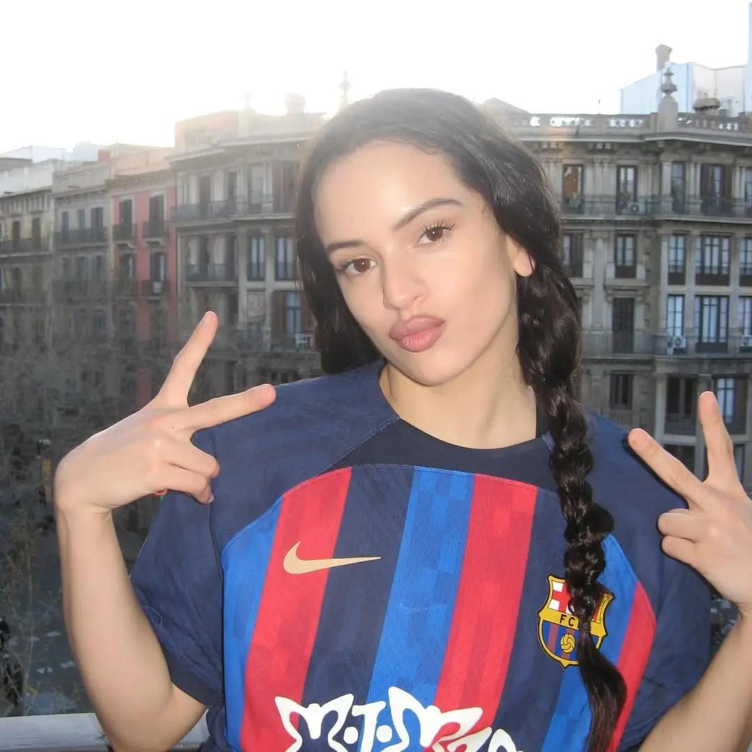 Young woman on a balcony posing for a photo in a Barcelona FC jersey, making peace signs with both hands.