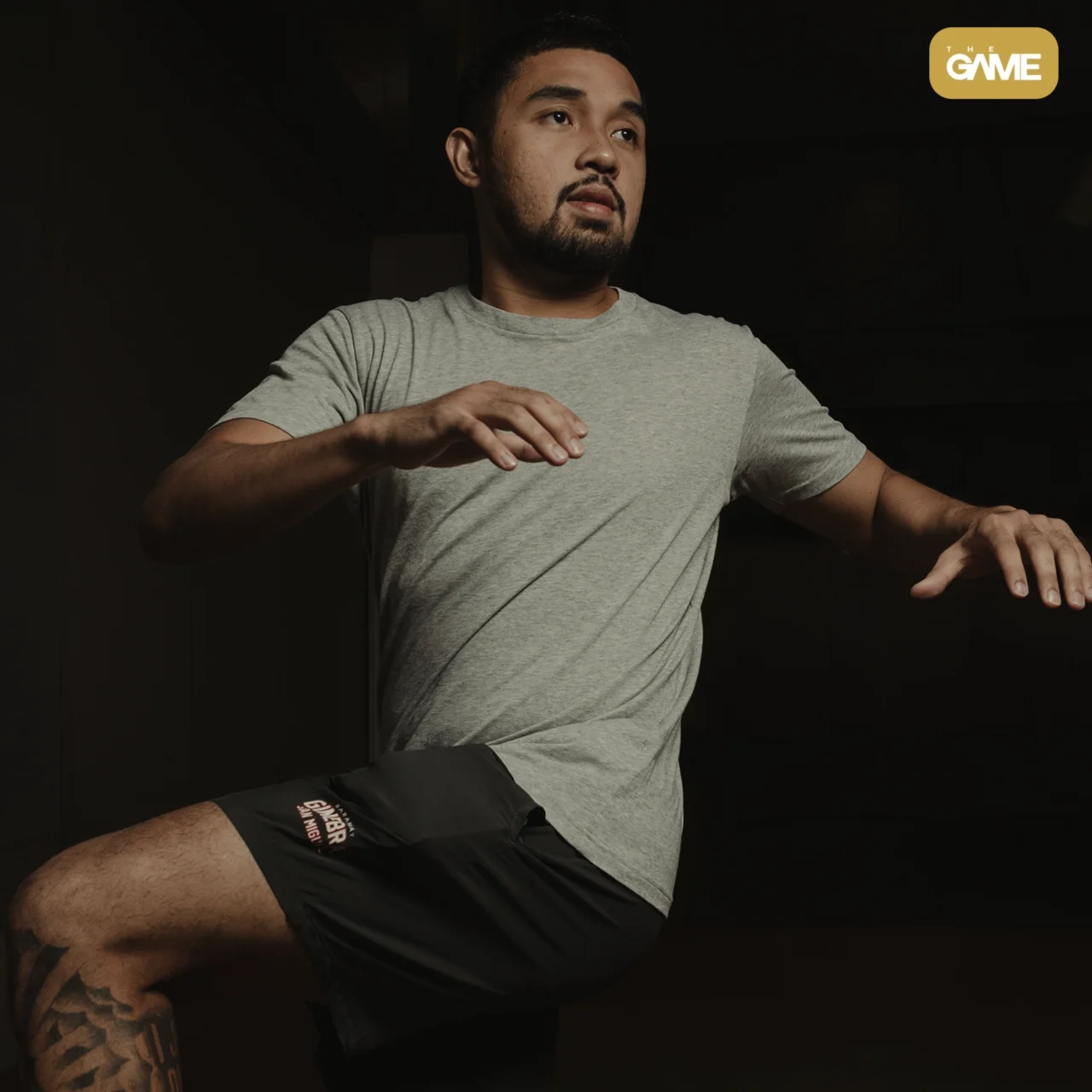 Man in a gray T-shirt demonstrating a mid-dance pose in a dark room, with The Game logo in the upper-right corner.
