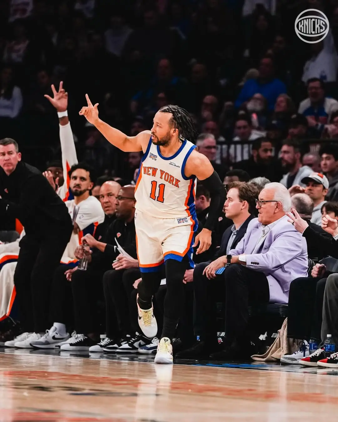 Saveclipapp_653661634_18574282681010608_5287351266810767189_n - the game Basketball player in Knicks white jersey with orange and blue trim (#11) gestures with one arm raised on the court as crowd watches from the stands and bench nearby.
