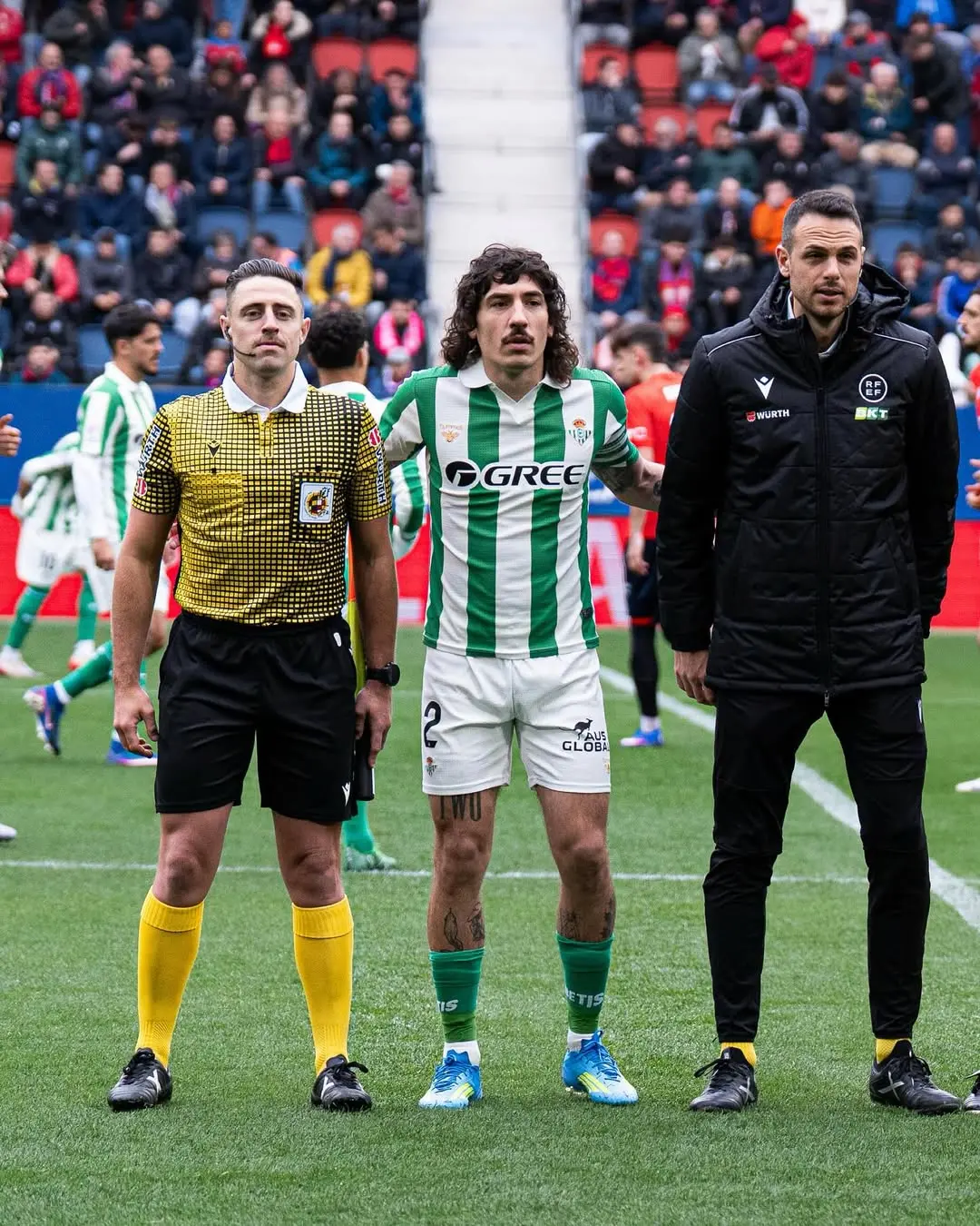 Three people stand on a soccer field before kickoff: a referee in a yellow patterned shirt, a green-and-white striped player in the middle, and a staff member in a black jacket on the right, with spectators in the stands behind them.