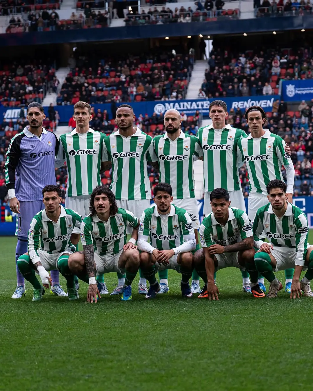 Professional soccer team posing for a group photo on the field in green-and-white striped jerseys, with stadium spectators in the background.