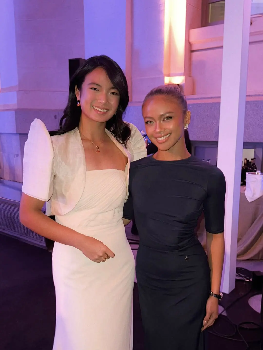 Two women posing together at a formal event; one in a white gown with puff sleeves, the other in a dark dress, both smiling.