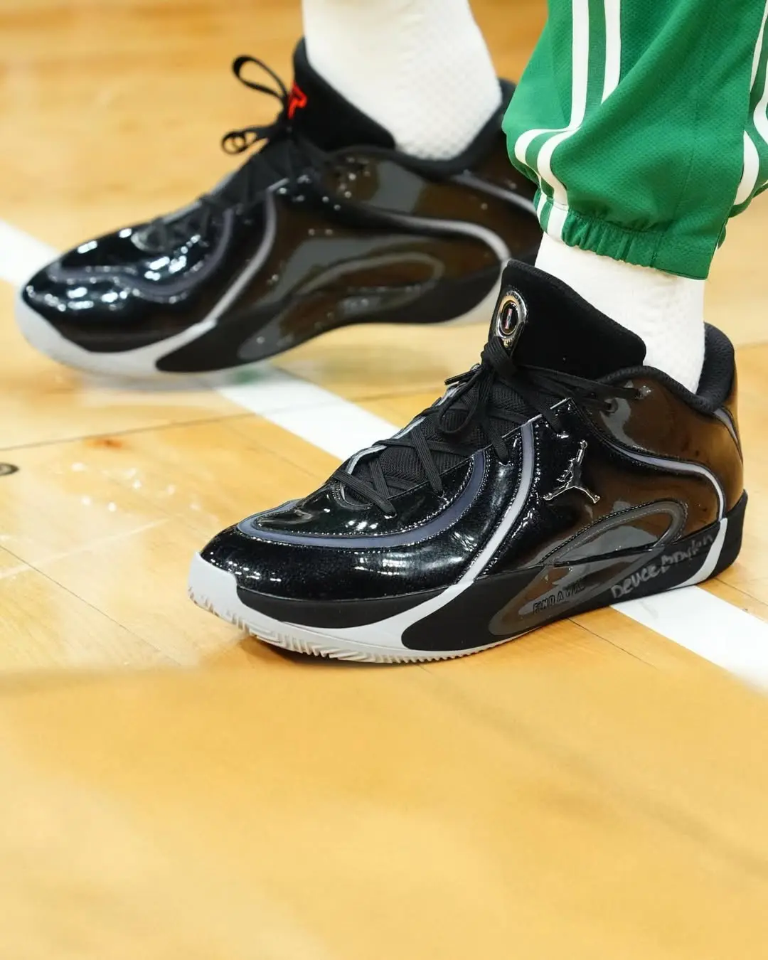 Black glossy basketball sneakers with white soles on a gym floor, worn with white socks and green athletic pants.