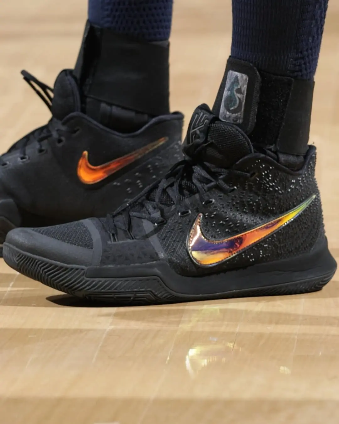 Black basketball shoes with orange iridescent Nike swoosh on a wooden court floor, worn with dark socks and leg sleeves.