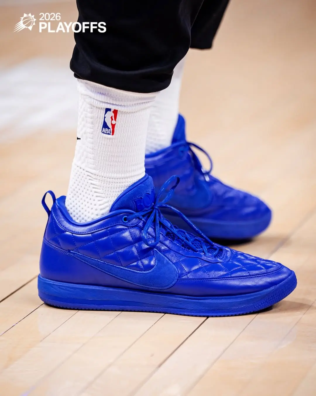 Close-up of bright blue basketball sneakers with white NBA logo socks on a wooden court. They appear ready for play, with '2026 PLAYOFFS' text in the corner.