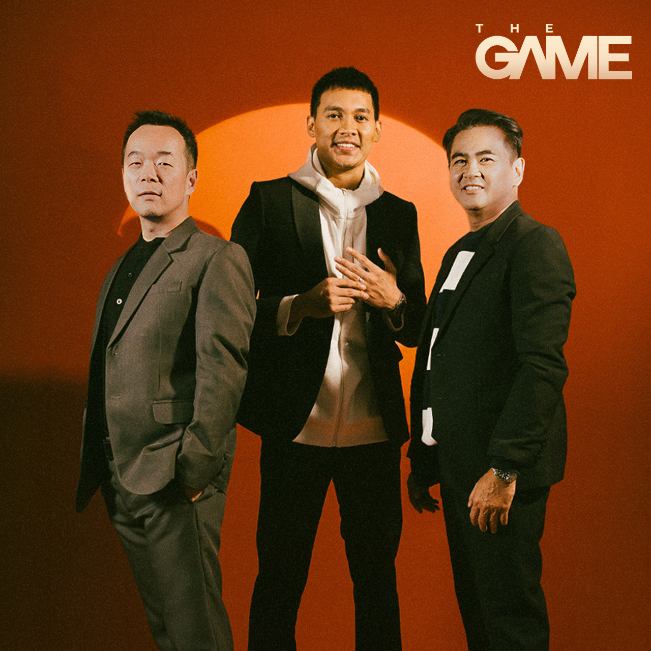 Three men in suits pose together against an orange backdrop with a pale orange circle behind them and THE GAME logo in the corner.