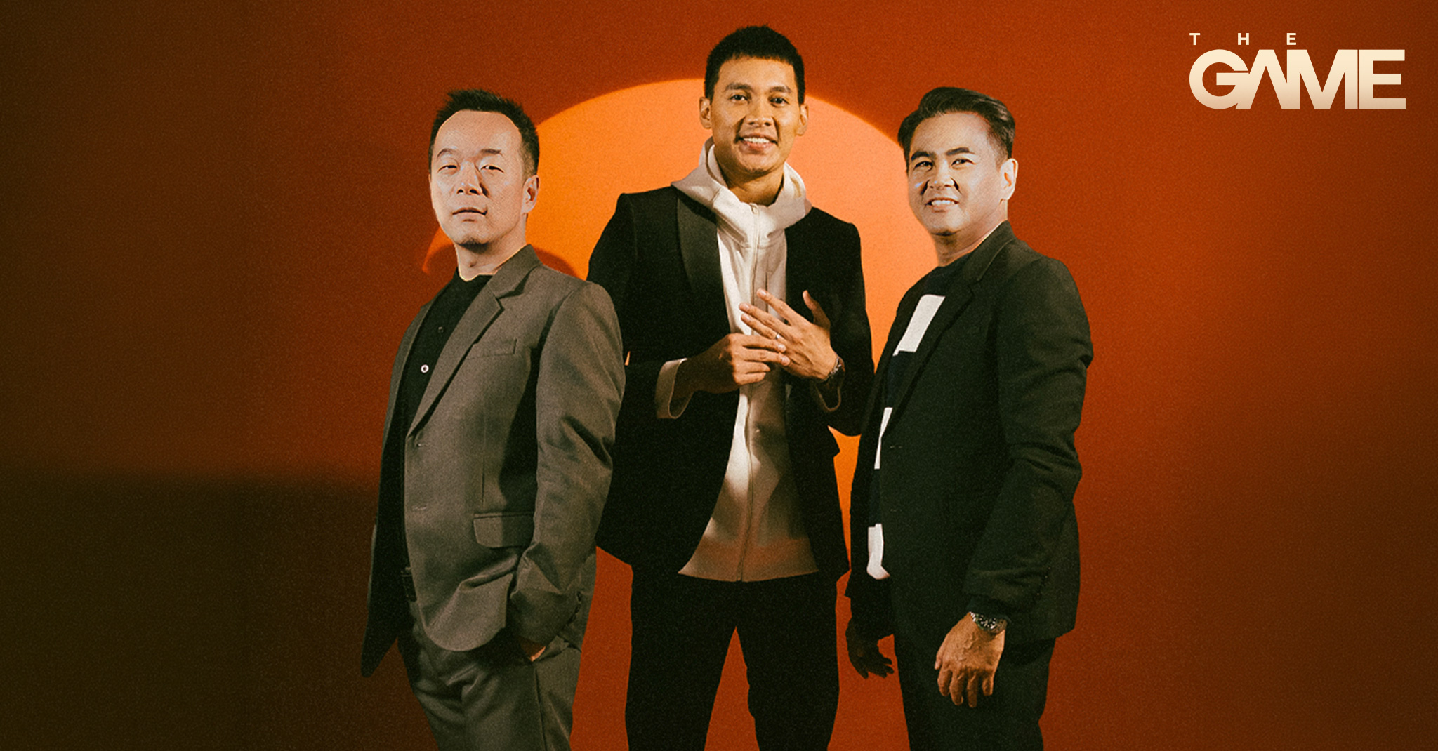 Three men in suits pose together against an orange backdrop with a pale orange circle behind them and THE GAME logo in the corner.