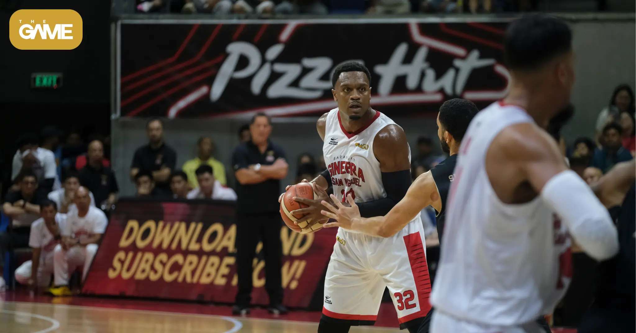 The game watermark banner wide 42 - the game Basketball player in white and red jersey (#32) grips the ball while defenders close in, with a crowd and a Pizza Hut sign in the background.
