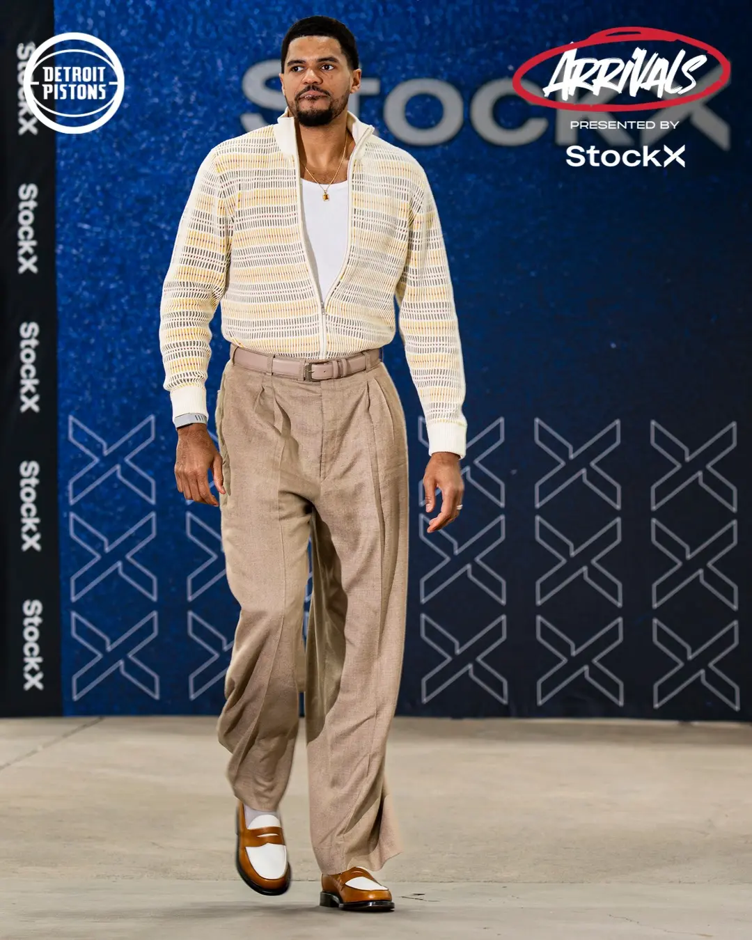 Male model walking a fashion runway in beige pleated trousers, white shirt, and striped cardigan at a StockX Arrivals event. (Runway scene)