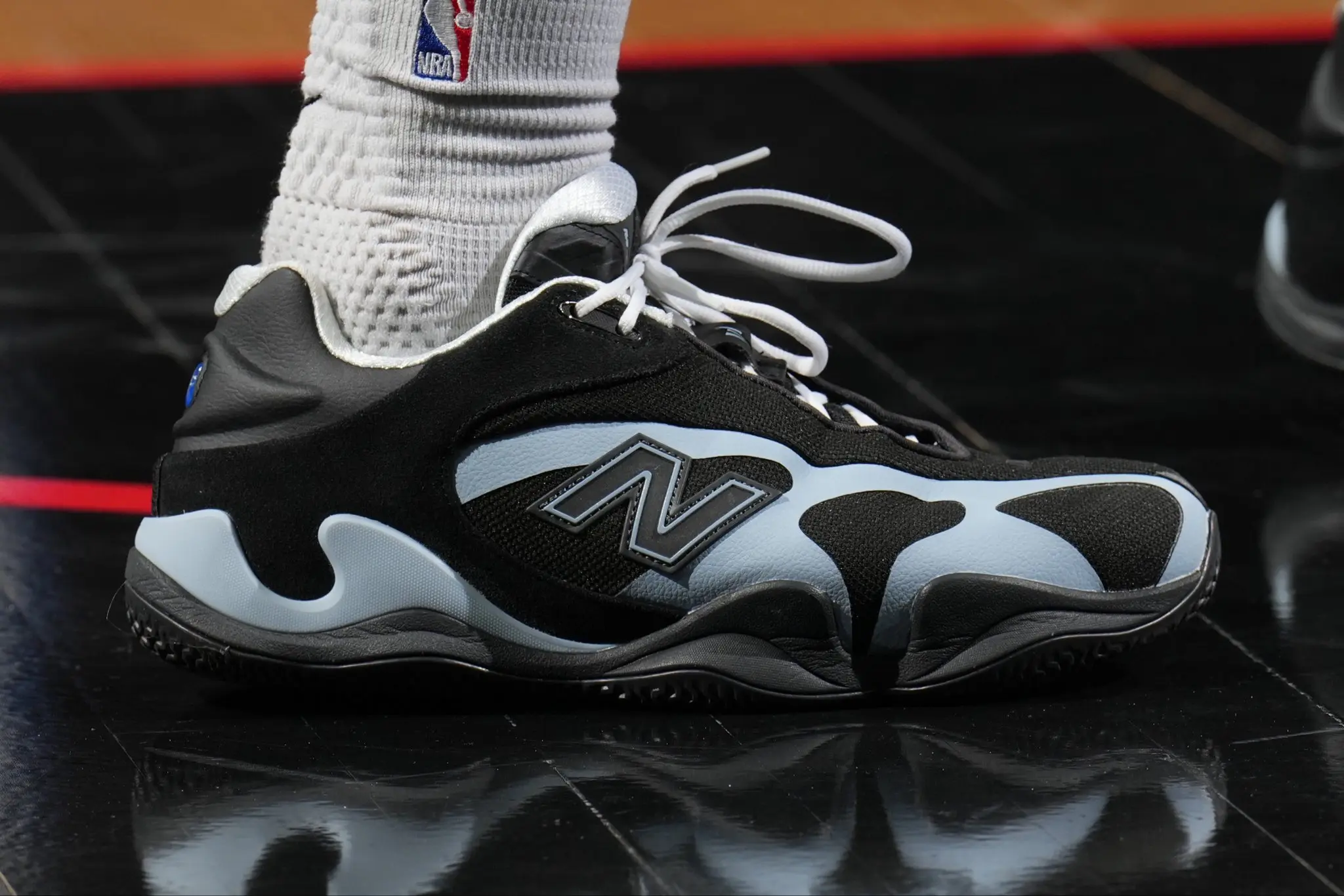 Close-up of a black, gray, and white athletic sneaker with white laces, worn with a textured white sock on a glossy surface (NBA logo visible).
