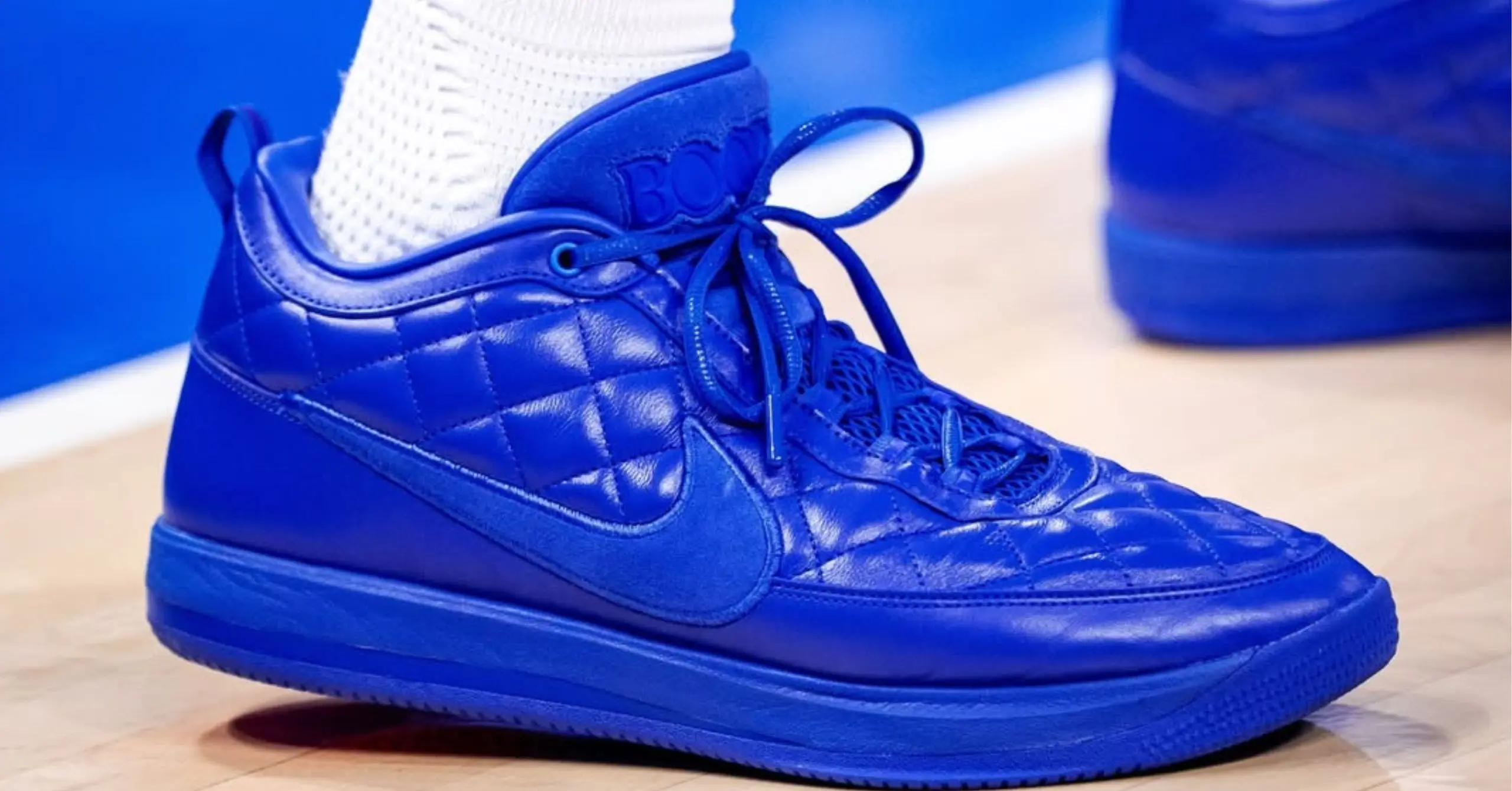 Blue quilted high-top sneaker with laces, resting on a wooden floor next to a blue object in the background