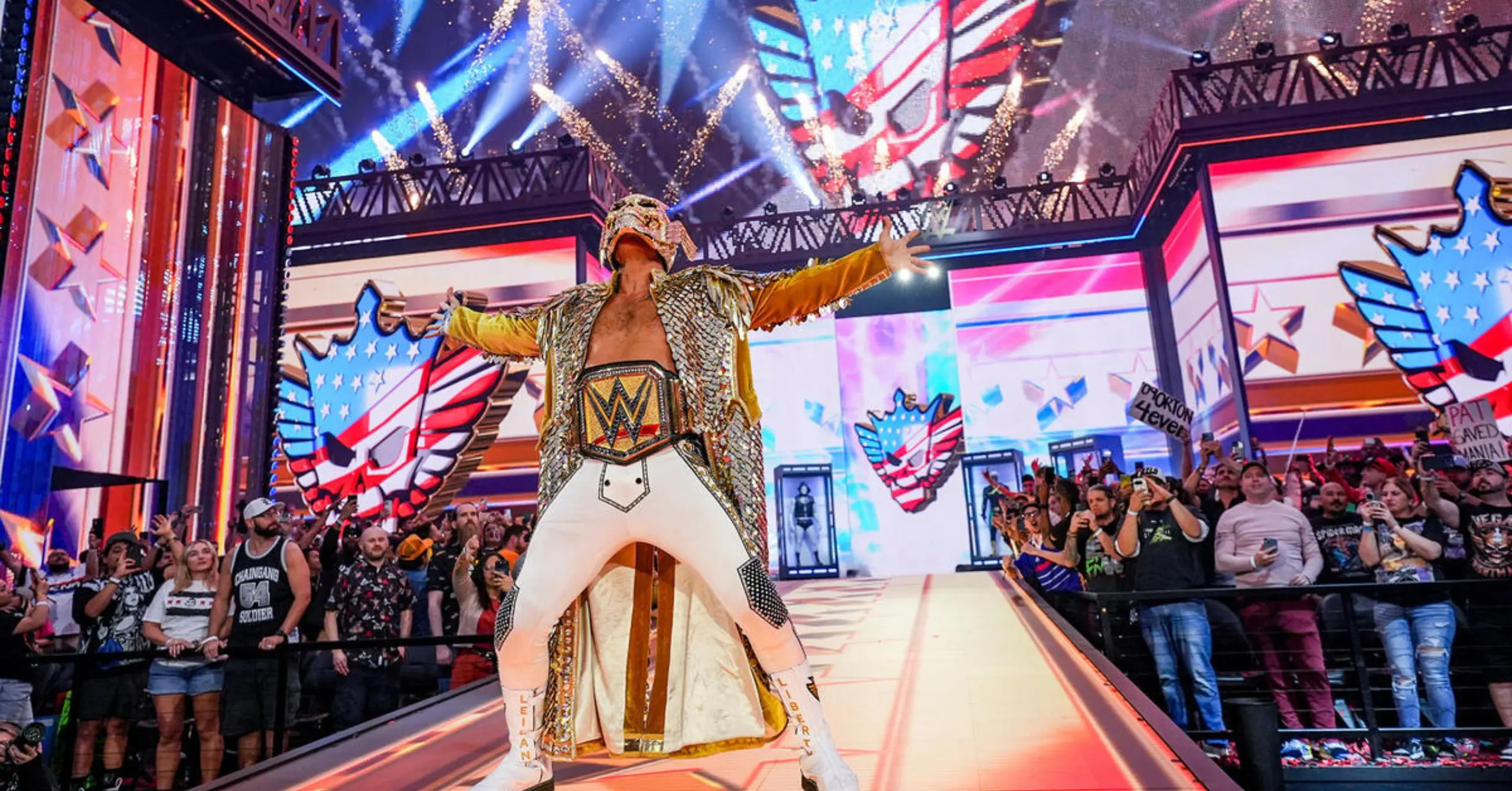 WWE wrestler in a gold cape and championship belt stands on a brightly lit stage with arms outstretched as fans cheer and hold signs nearby.