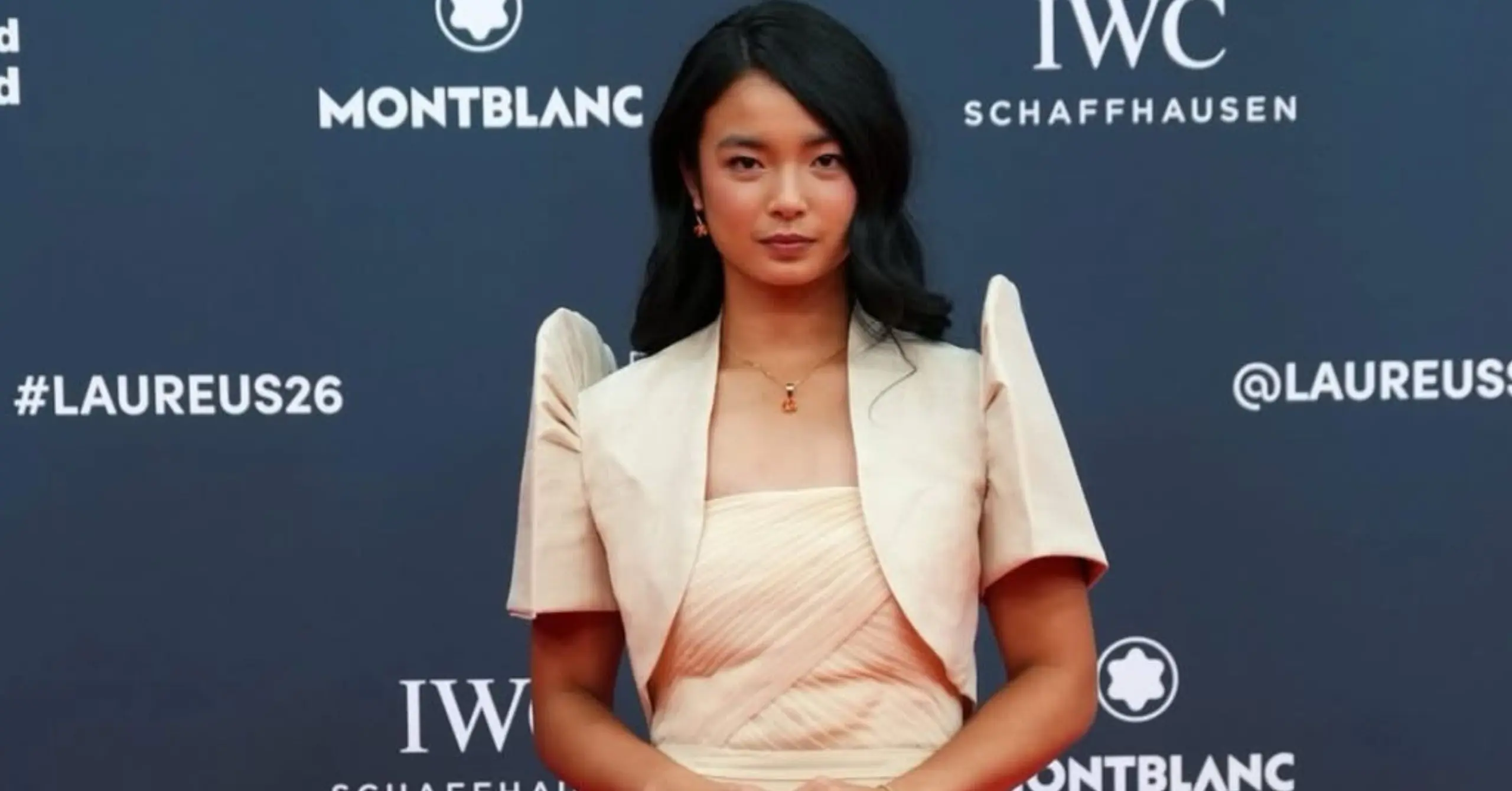 Woman posing on a blue backdrop with sponsor logos (Montblanc, IWC) wearing a pale peach gown and necklace at a Laureus event.