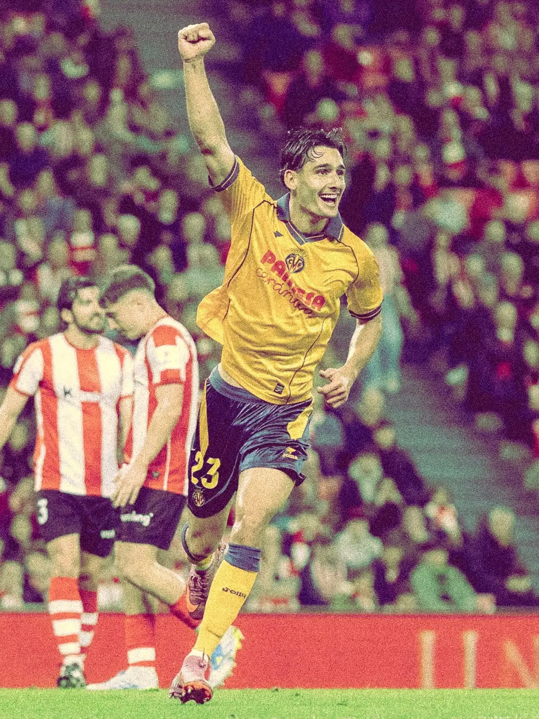 Soccer player in a yellow kit with number 23 raises fist in celebration on the pitch, crowd in the background.