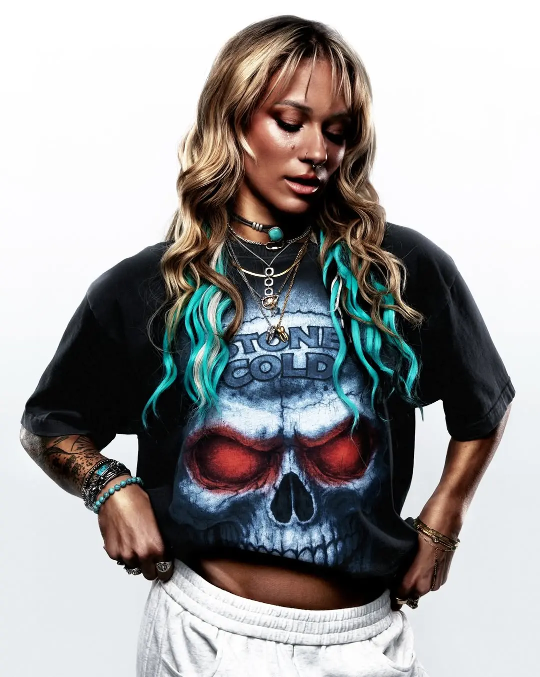 Woman with wavy blonde hair and teal tips wearing a black graphic t-shirt featuring a blue skull with red eye sockets and a white background.