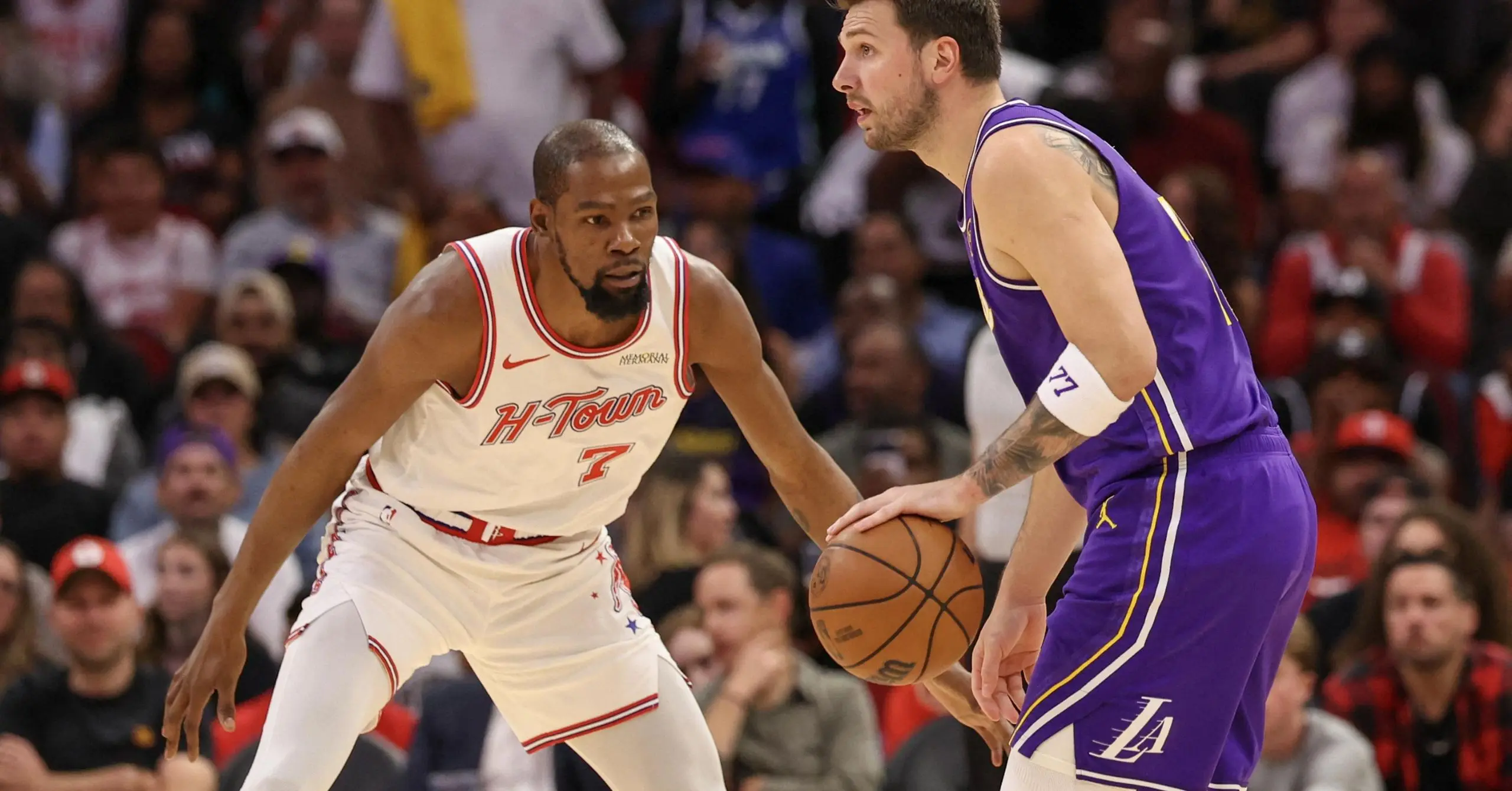 Houston Rockets guard in white jersey (#7) defends a Lakers player in purple who is dribbling the ball nearby at a crowded game stadium.