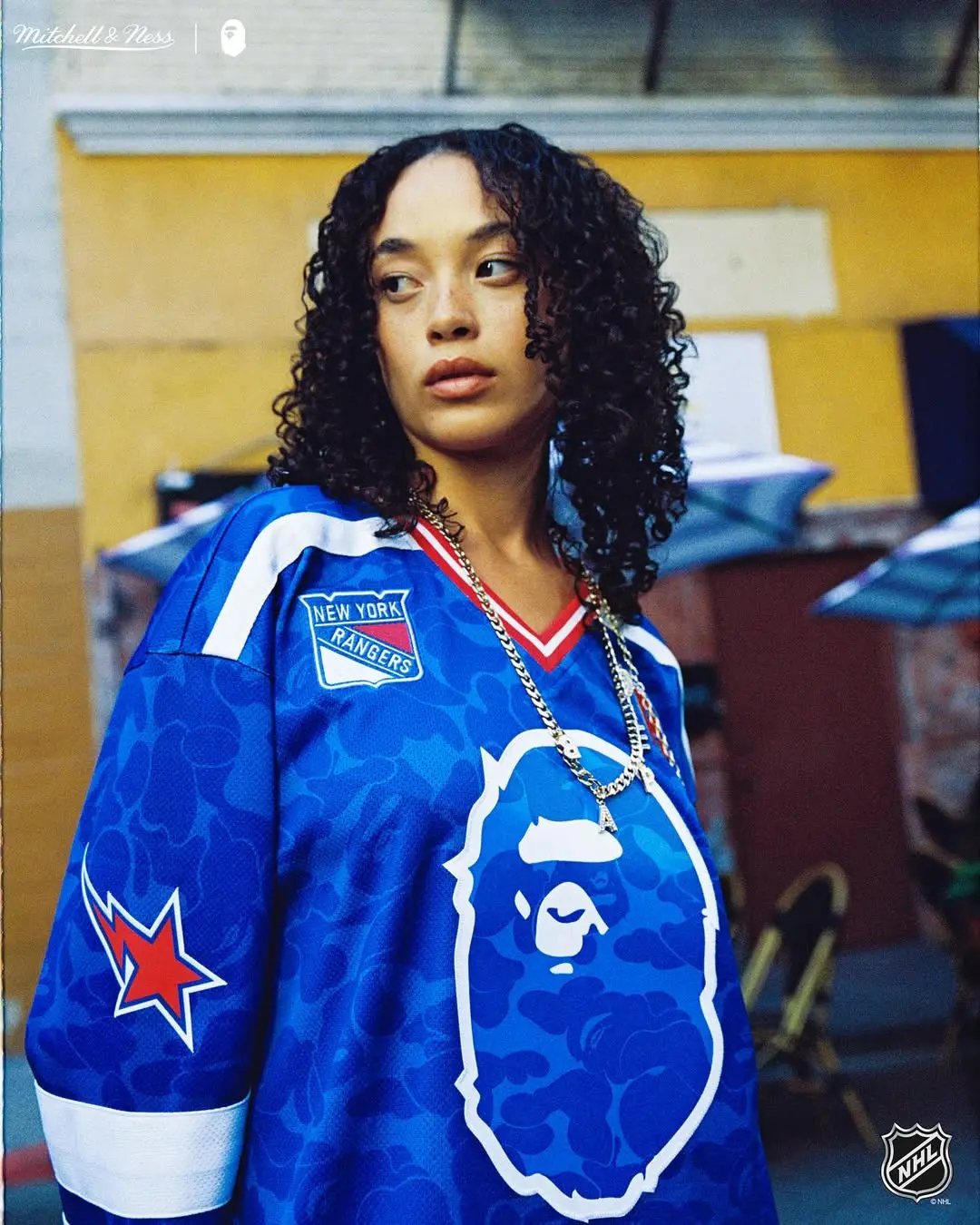 Portrait of a woman with curly hair wearing a blue camouflage hockey jersey featuring a large ape logo and a New York Rangers patch, outdoors.