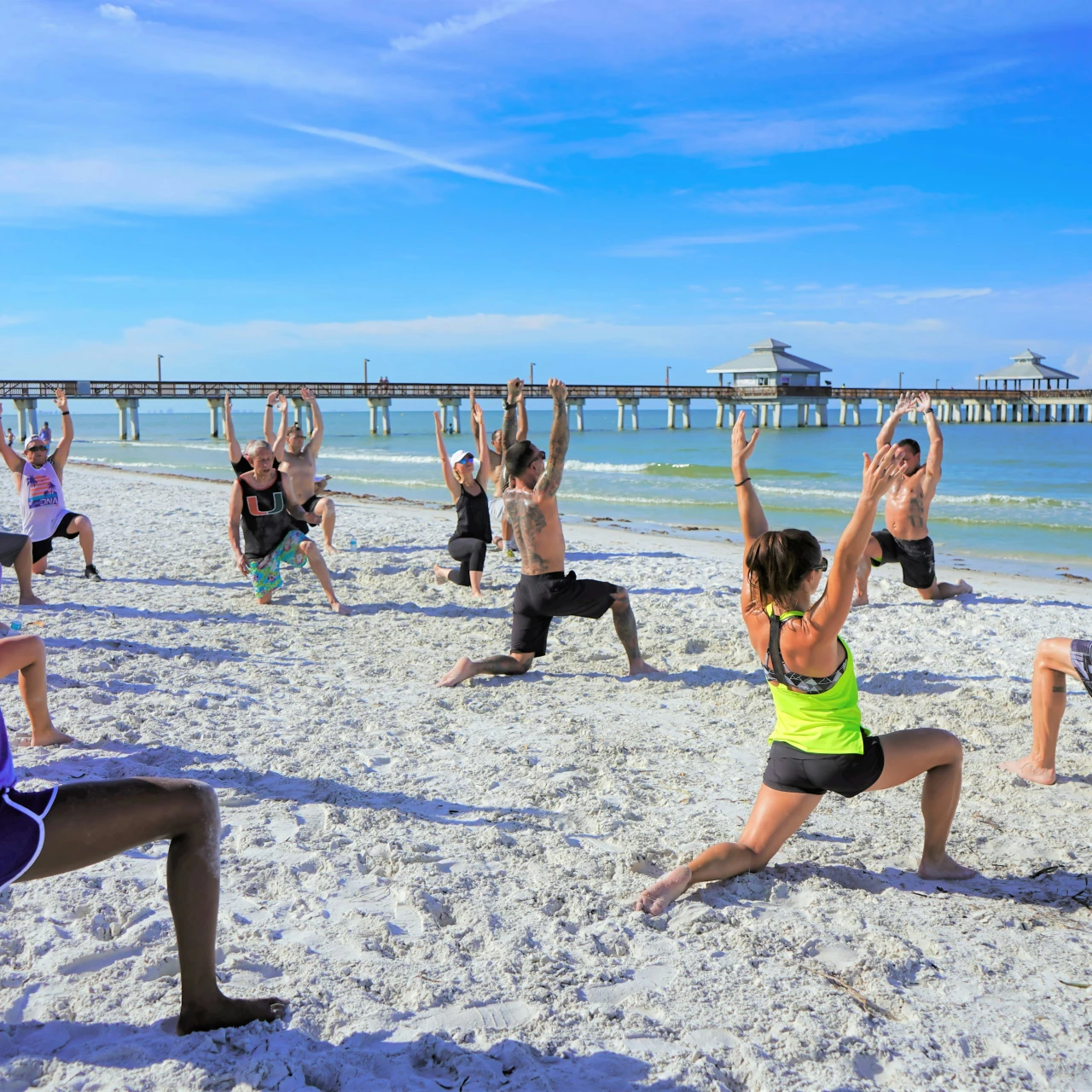 Group of people doing a yoga session on a sandy beach with a wooden pier and ocean in the background.